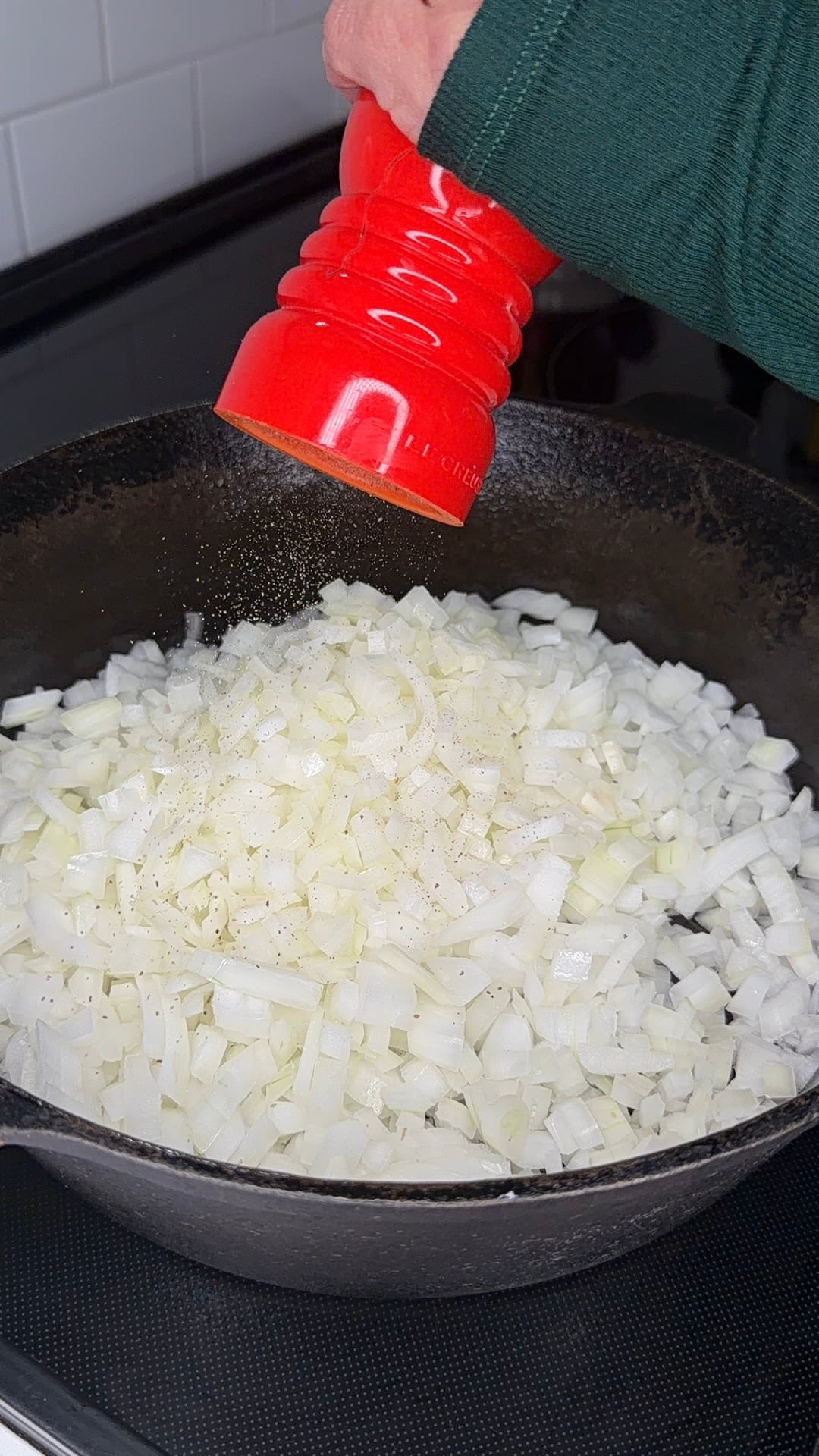 A hand holds a red pepper grinder over a cast iron skillet filled with chopped onions, about to season them. White tile backsplash and stovetop are visible in the background.