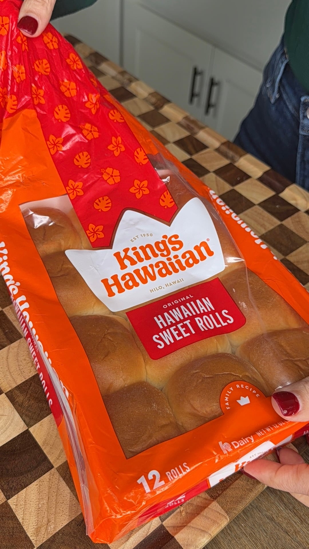 A person with red-painted nails opens a package of Kings Hawaiian Original Hawaiian Sweet Rolls on a checkered wooden countertop.