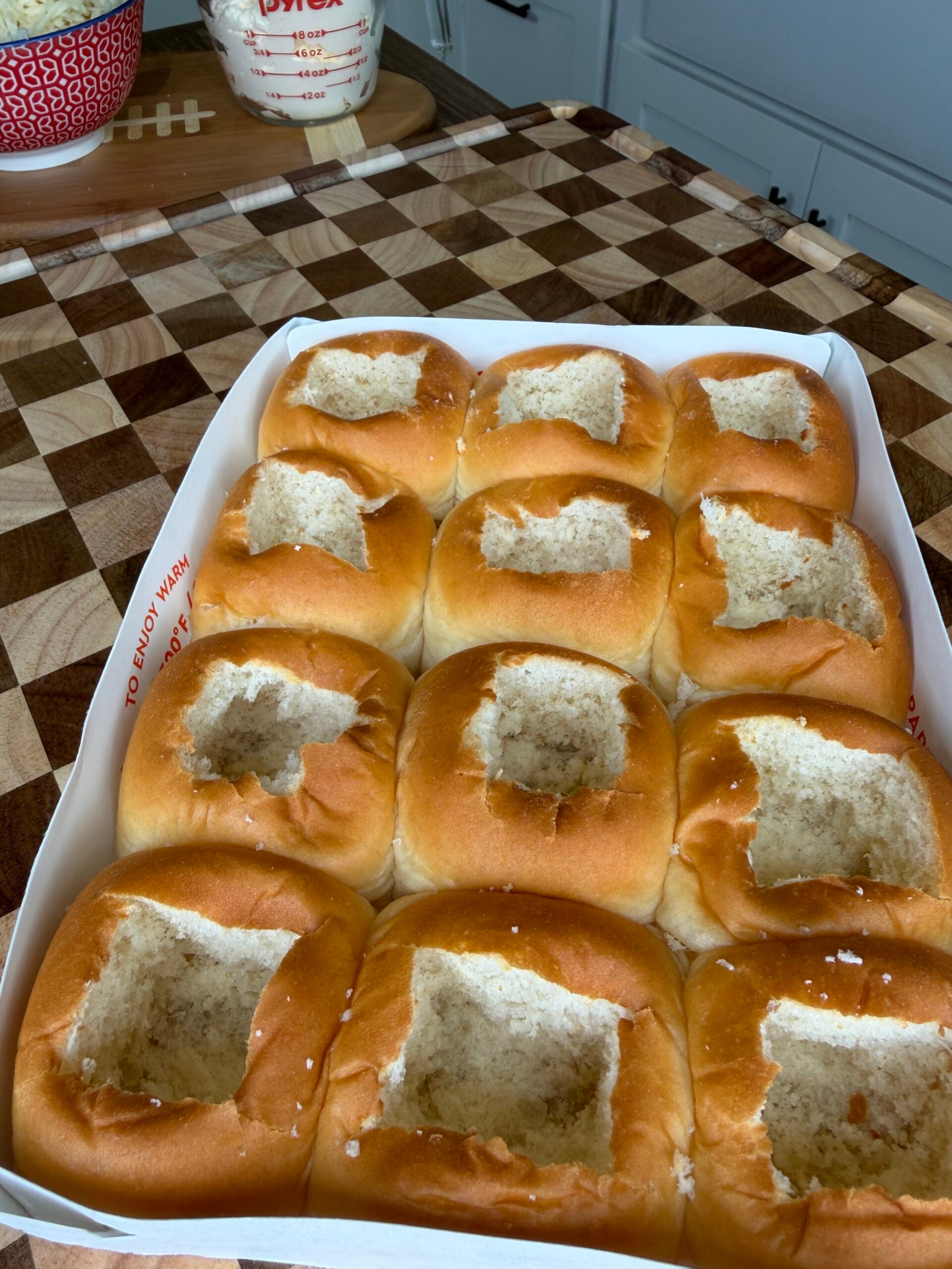 A tray of twelve soft dinner rolls with square holes cut out of their tops, set on a checkered wooden surface in a kitchen.