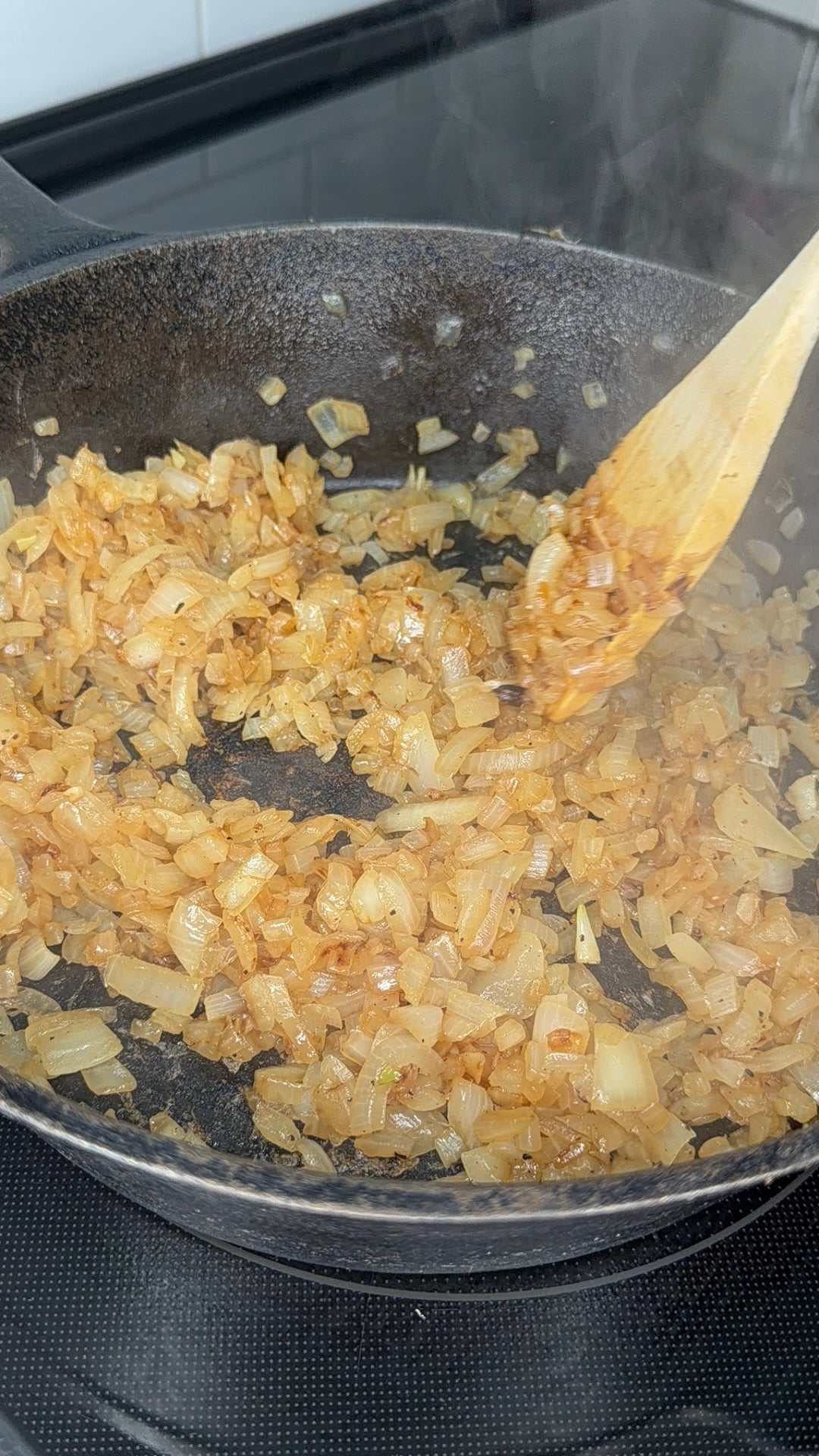 Chopped onions being sautéed in a black frying pan, stirred with a wooden spoon, starting to brown with visible steam rising.