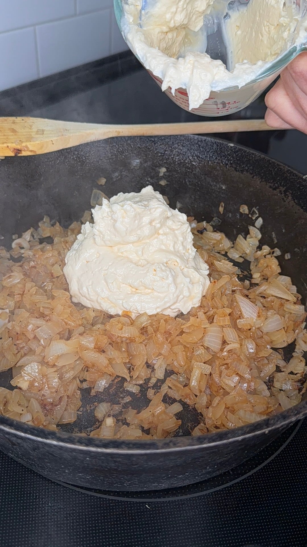 Chopped onions are sautéing in a black skillet, while a large dollop of creamy mixture is being added from a bowl held above the pan. Steam is rising from the hot onions.