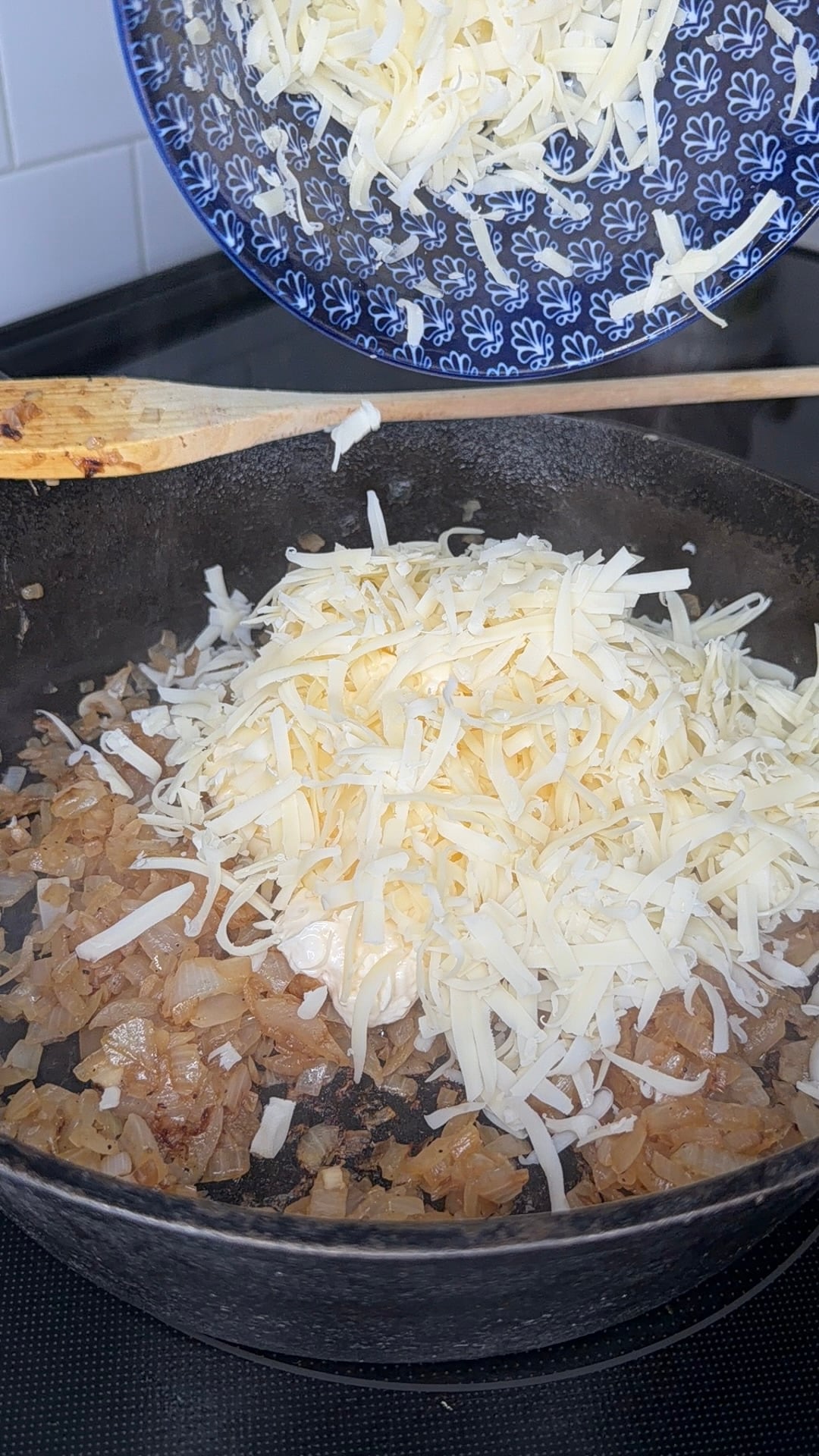 Grated cheese is being added from a patterned blue bowl to sautéed onions in a black cast iron skillet on a stovetop.