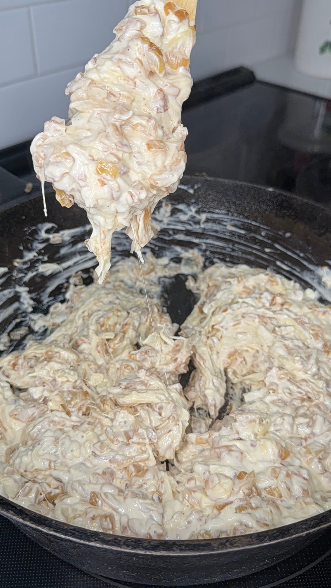 A close-up of a black skillet filled with a creamy, cheesy mixture being lifted with a wooden spoon, showing melted cheese stretching. White subway tiles are visible in the background.