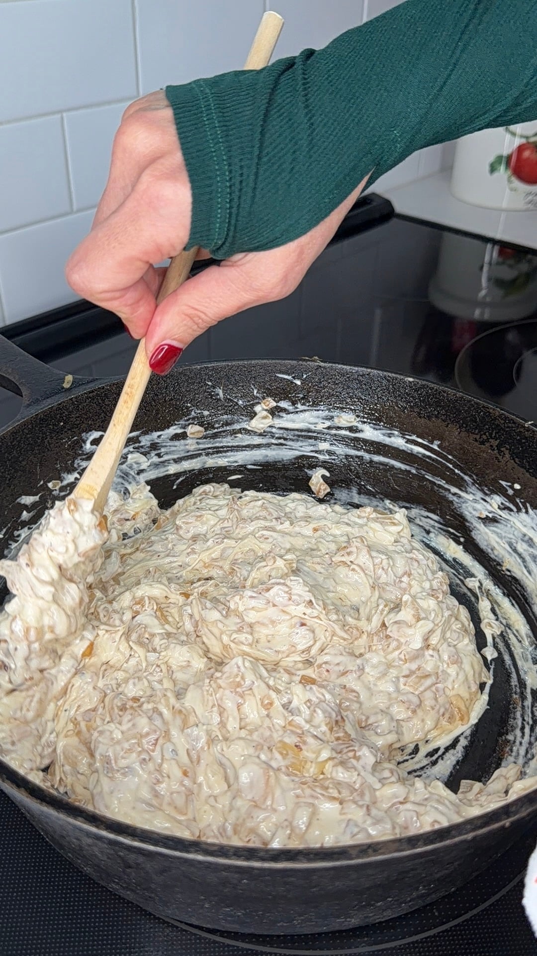 A hand with red nail polish stirs a creamy mixture in a black cast iron skillet on a stovetop using a wooden spoon.