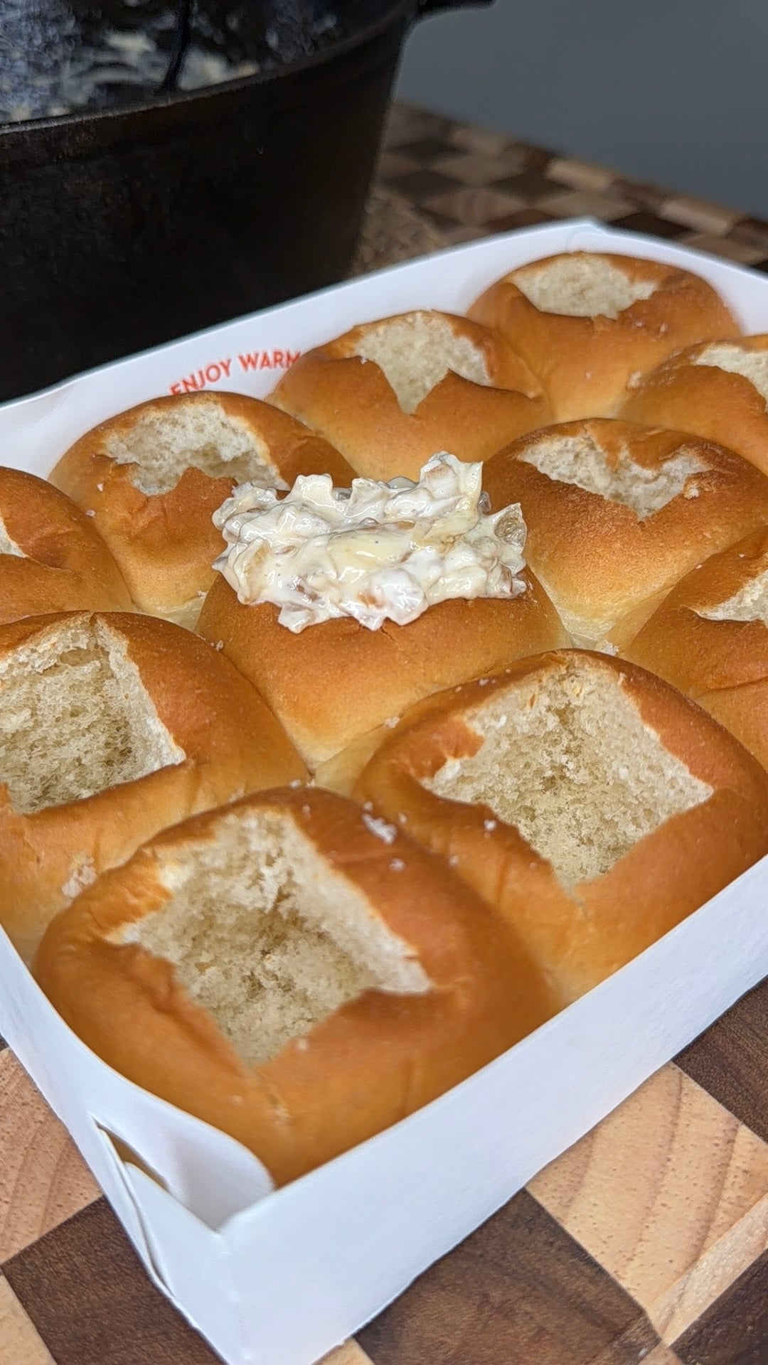 A box of nine pull-apart bread rolls, each with the top cut off. The center roll is filled with a creamy dip, while the others are hollowed out. The box sits on a wooden surface.