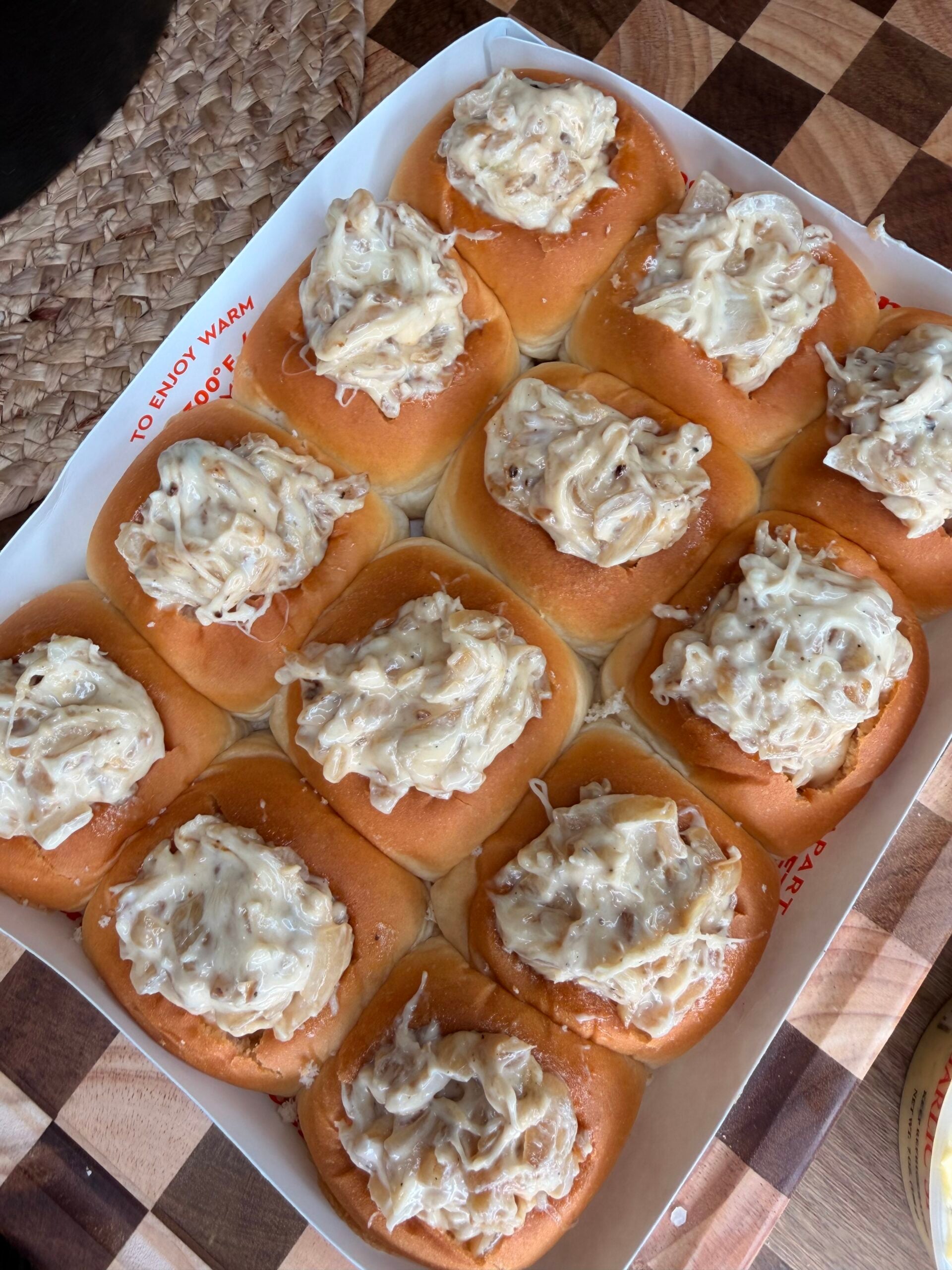 A tray of twelve slider buns topped with a creamy shredded chicken mixture sits on a checkered wooden surface next to a woven placemat.