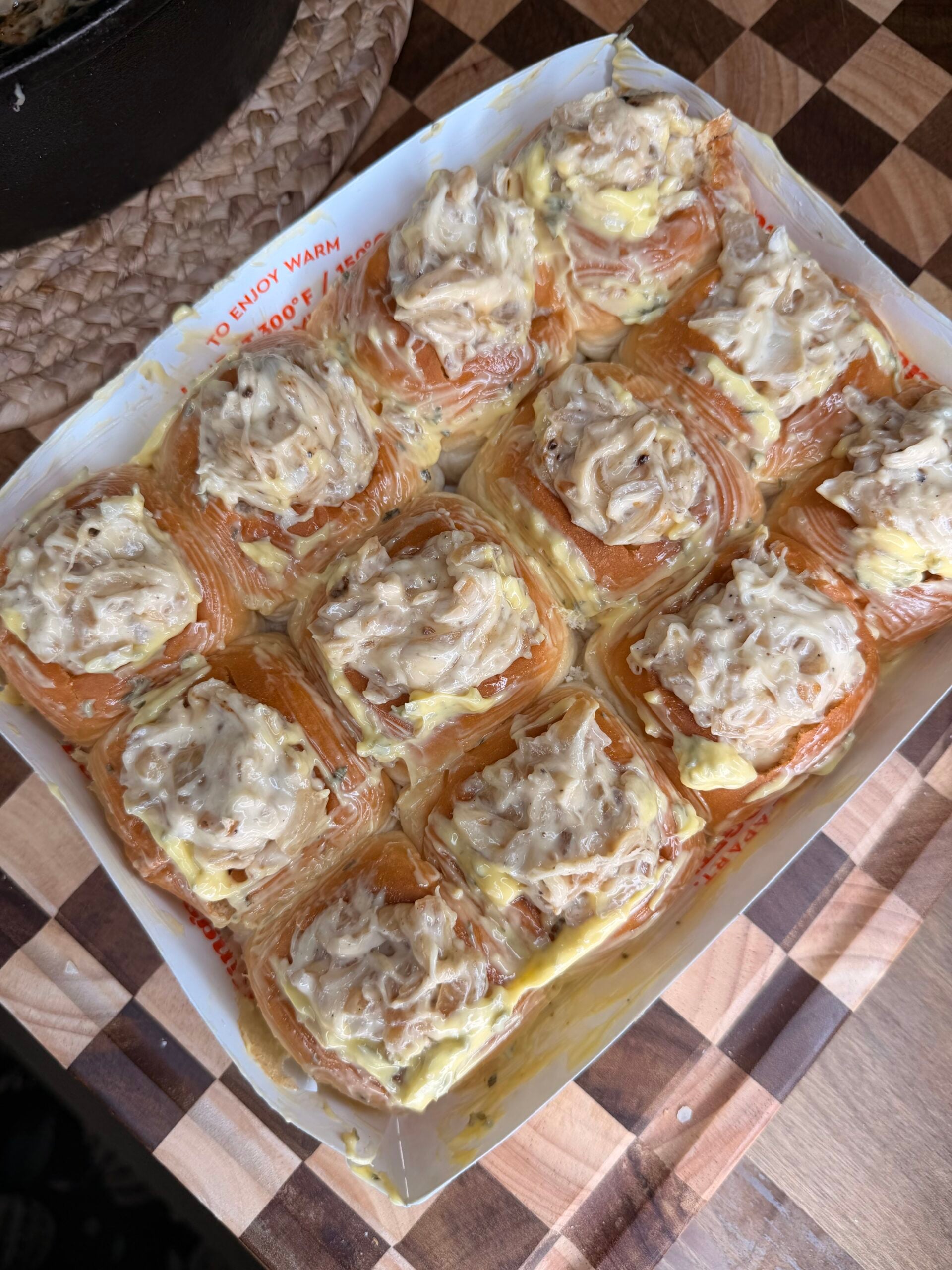 A tray of golden, flaky rolls topped with a creamy, chunky spread, resting on a patterned wooden surface.