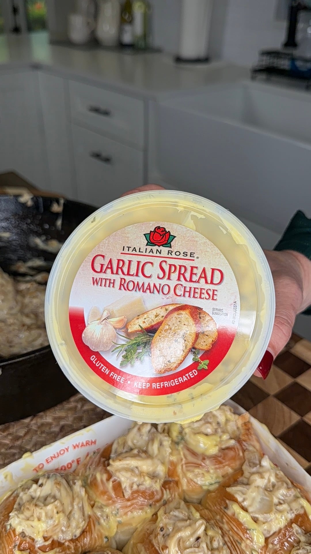 A hand holds a tub labeled Italian Rose Garlic Spread with Romano Cheese above pastries topped with the spread, in a bright kitchen setting.