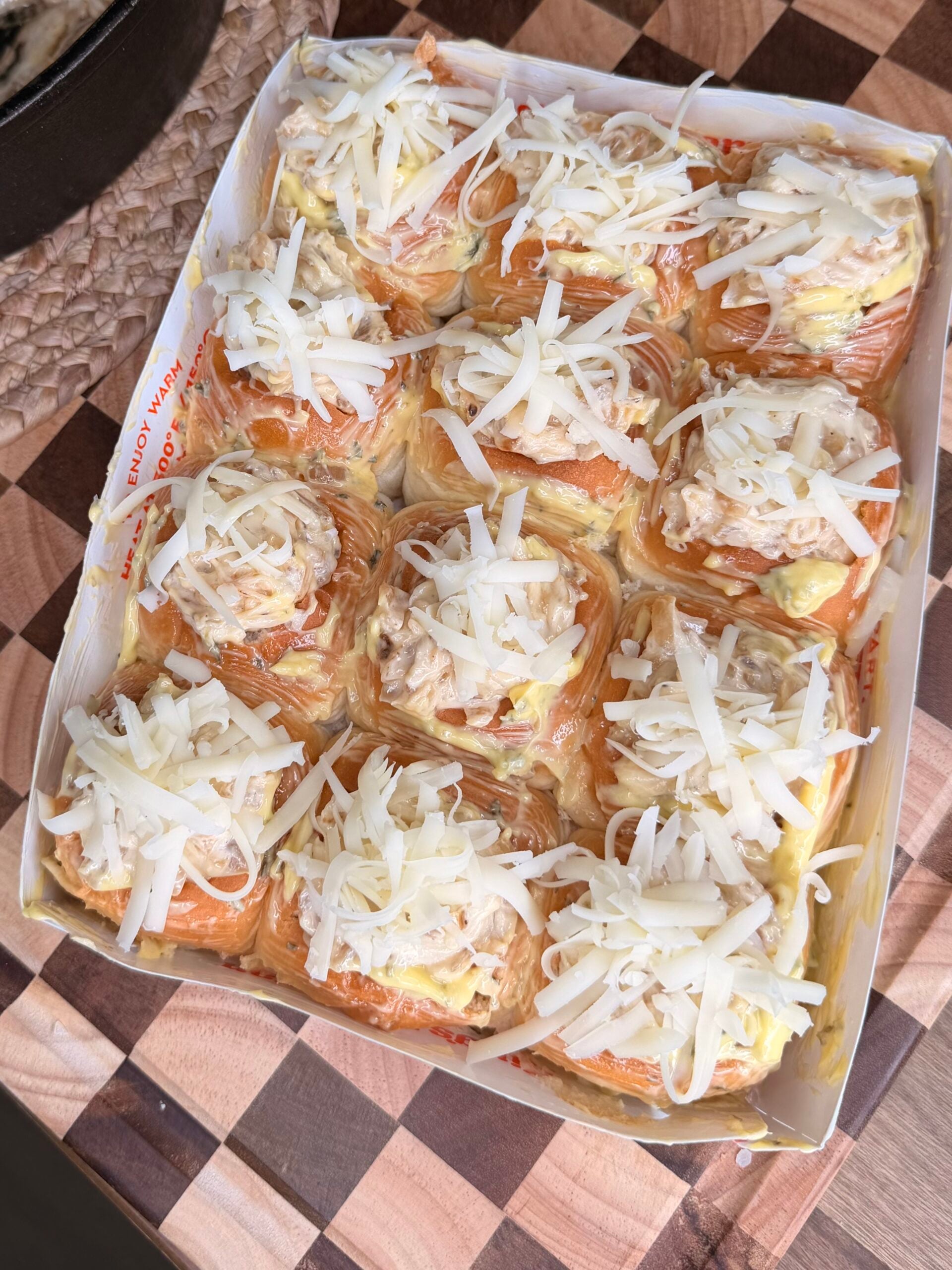 A tray of bread rolls topped with shredded cheese and a creamy mixture, arranged on a wooden cutting board.