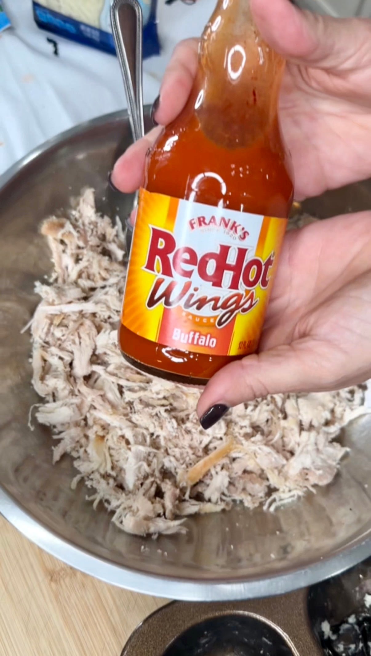 A hand holding a bottle of Franks RedHot Buffalo Wings sauce over a metal mixing bowl filled with shredded chicken.
