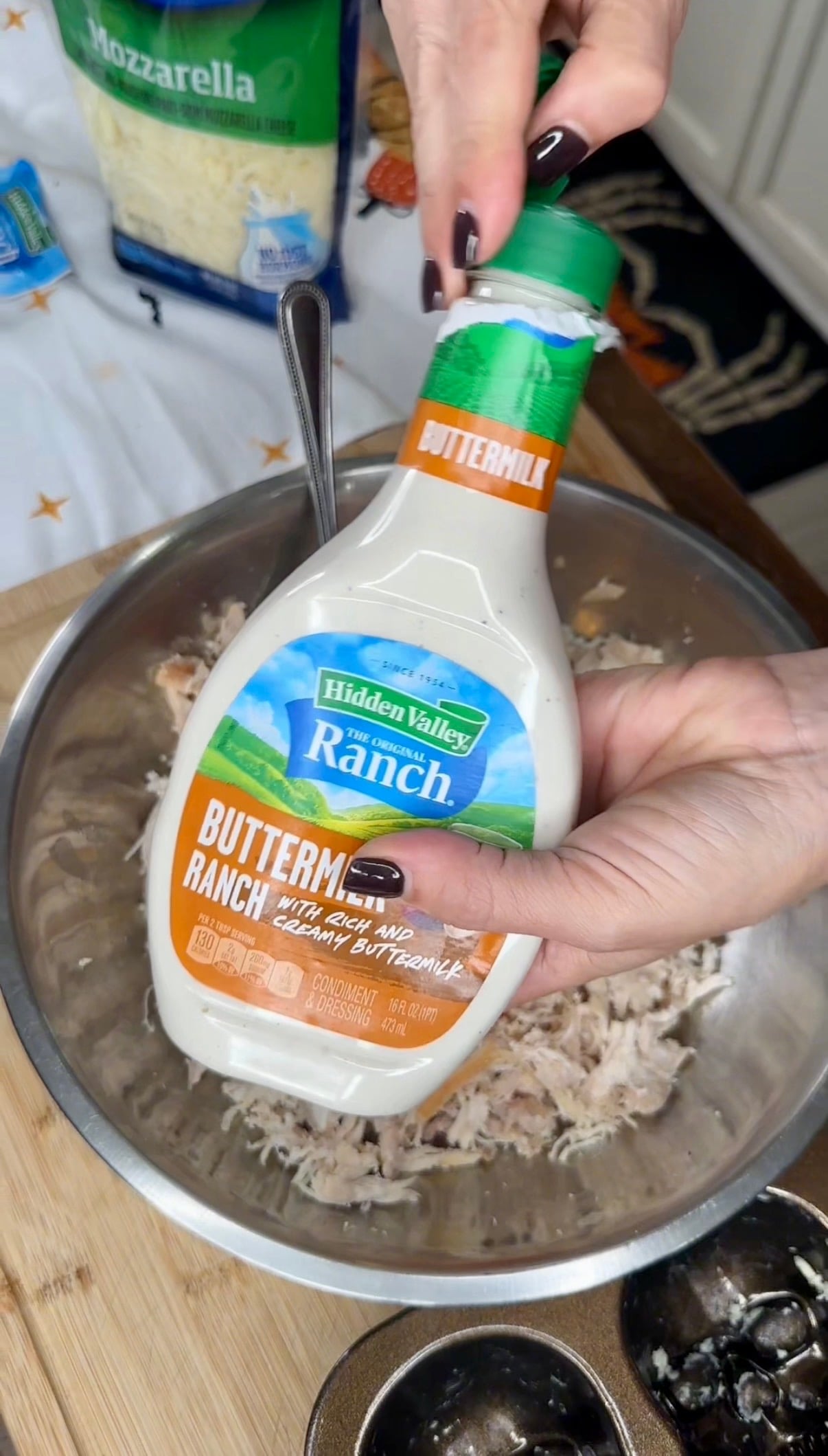 A person pours a bottle of Hidden Valley Buttermilk Ranch dressing into a metal bowl containing shredded chicken.