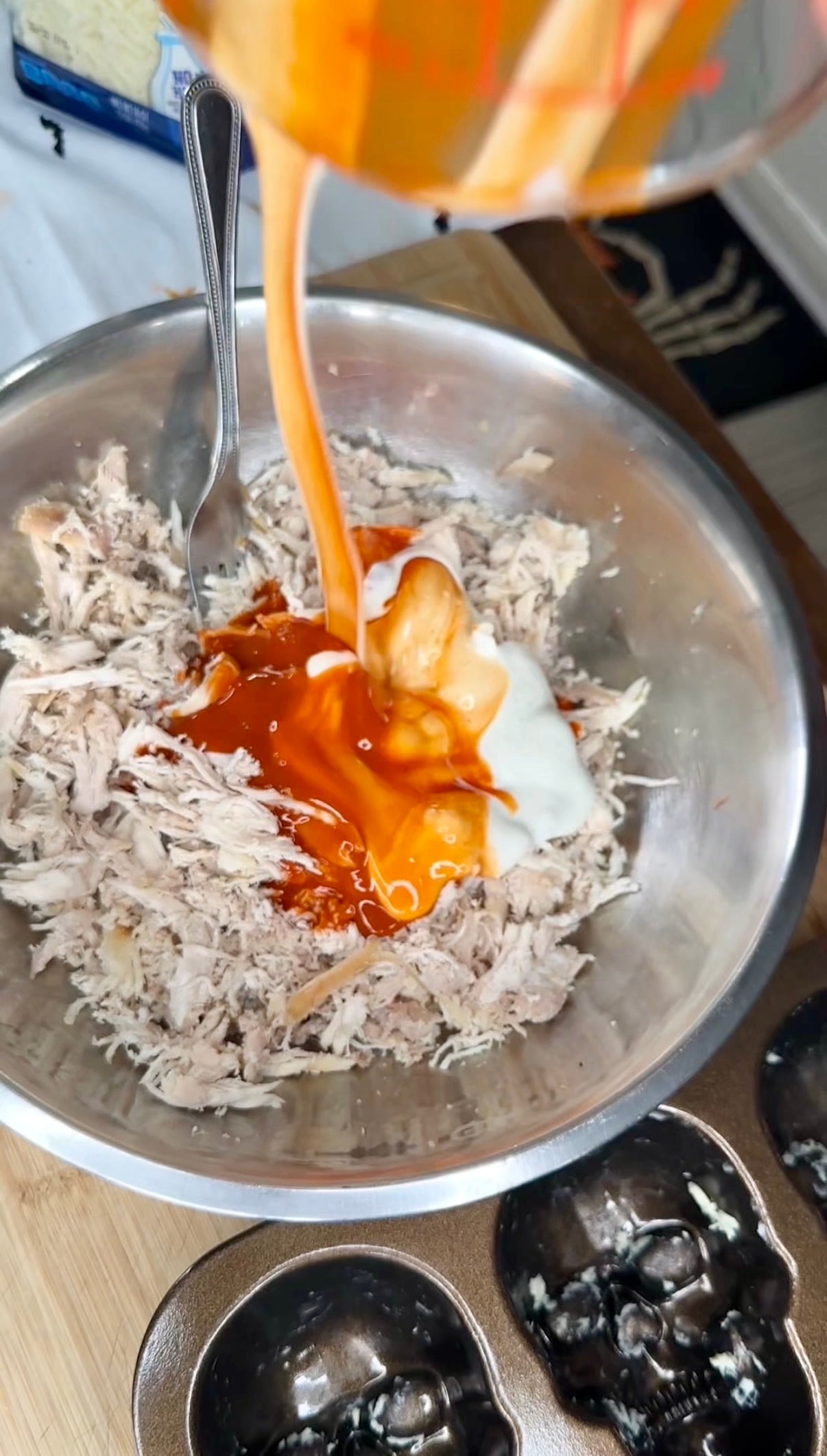 A measuring cup pours orange buffalo sauce and white dressing into a metal bowl containing shredded chicken.