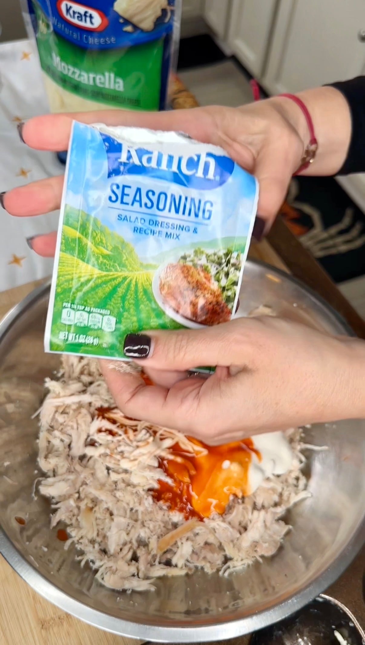 A pair of hands holding a packet of ranch seasoning mix over a metal mixing bowl containing shredded chicken, hot sauce, and sour cream.