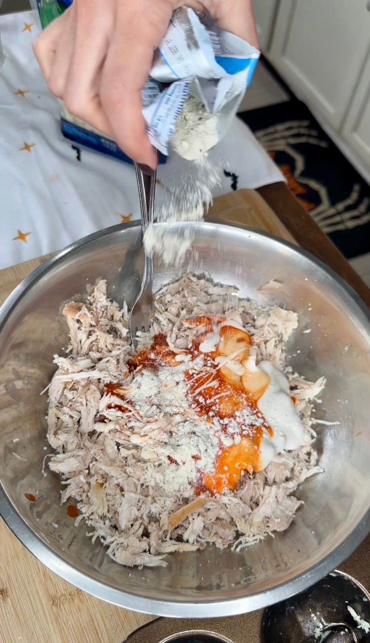 A hand pours a packet of seasoning into a metal bowl containing shredded chicken, hot sauce, and sour cream.