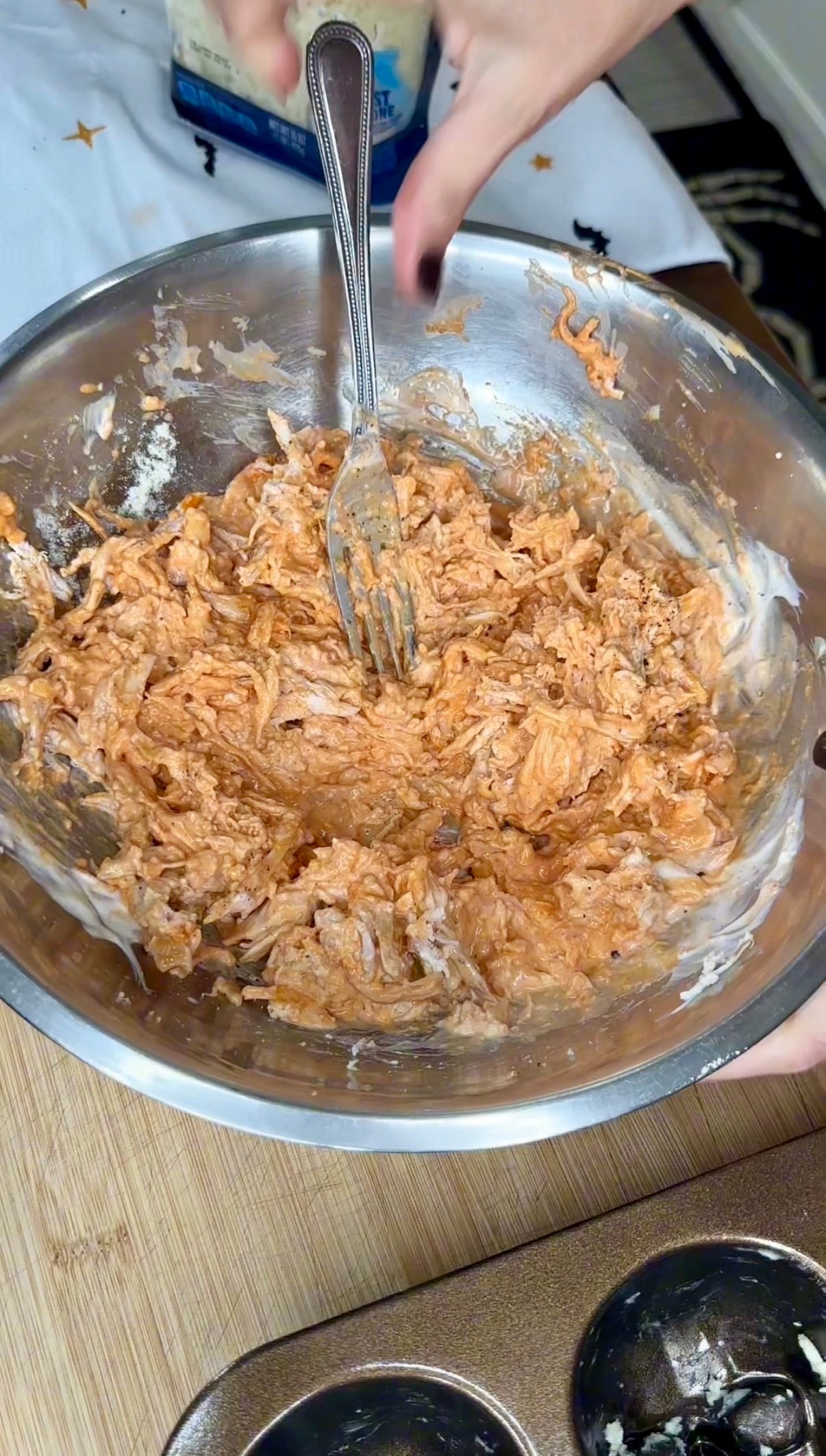 A metal bowl filled with shredded chicken mixed with a creamy orange sauce, being stirred by a fork.