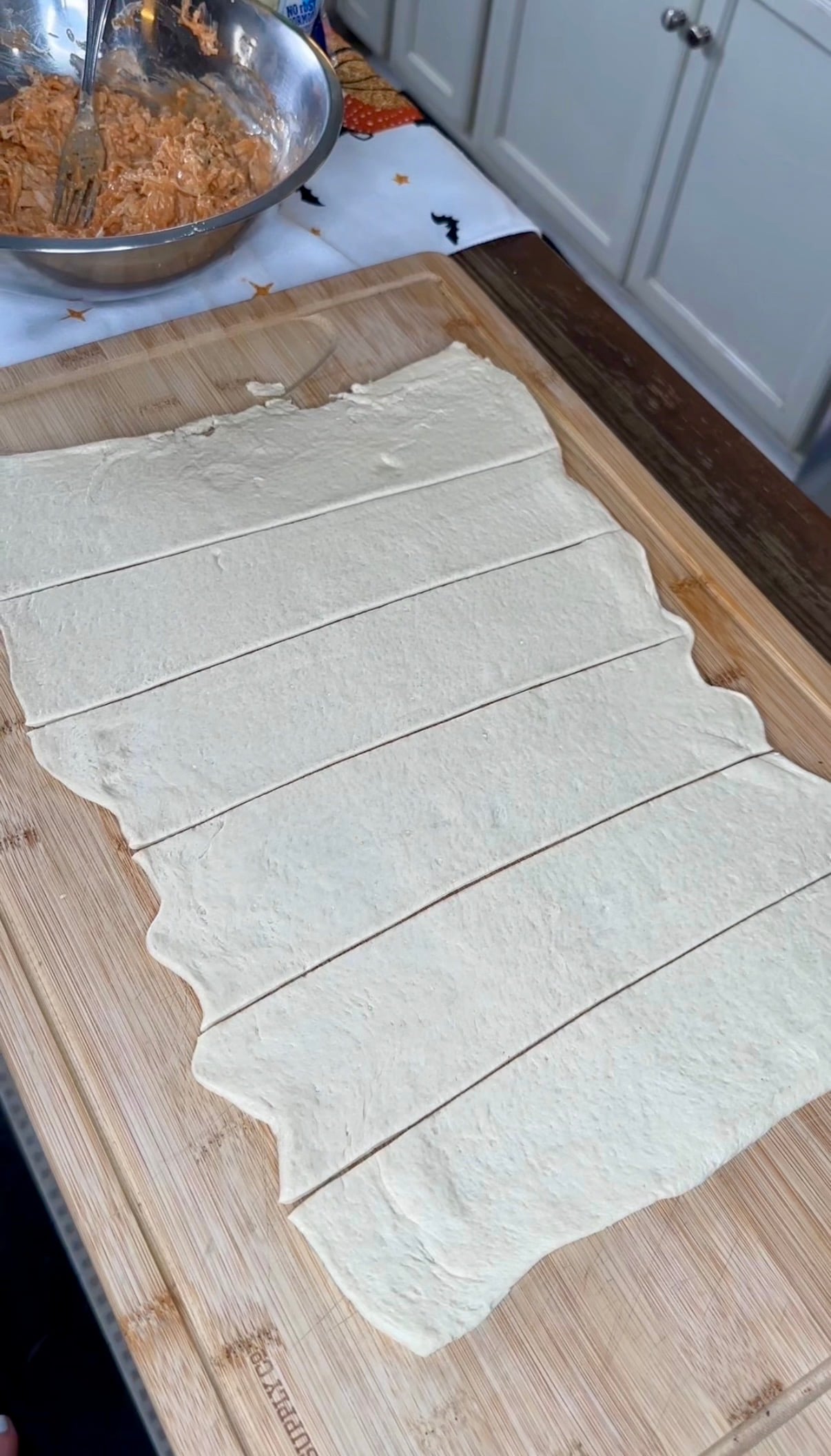 A rectangular sheet of raw dough sliced into strips on a wooden cutting board, with a bowl of shredded chicken mixture nearby.