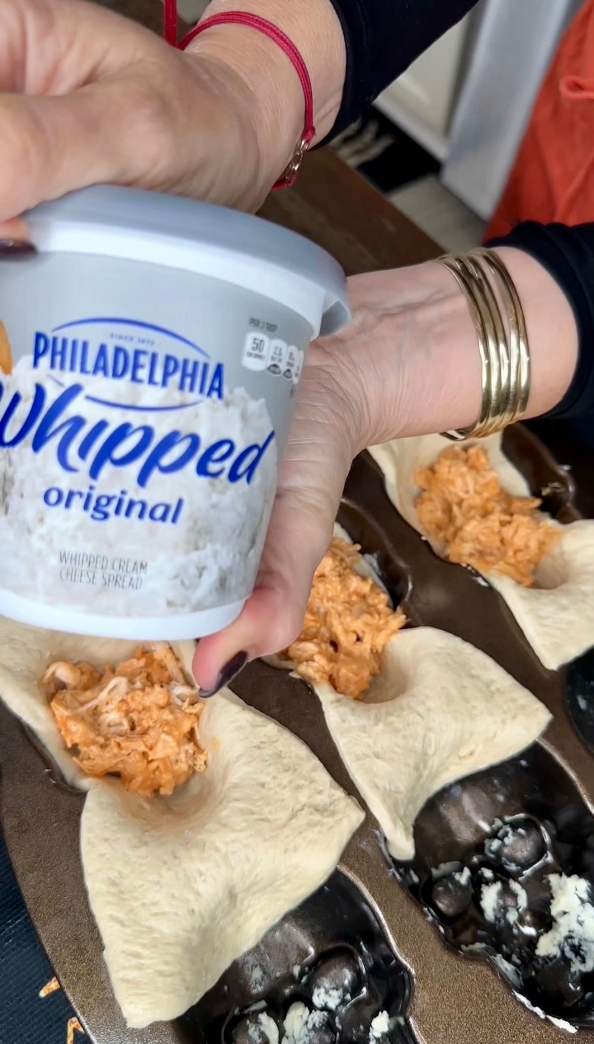 A hand holds a tub of Philadelphia Whipped cream cheese over a muffin tin containing scoops of buffalo chicken mixture inside dough shells.