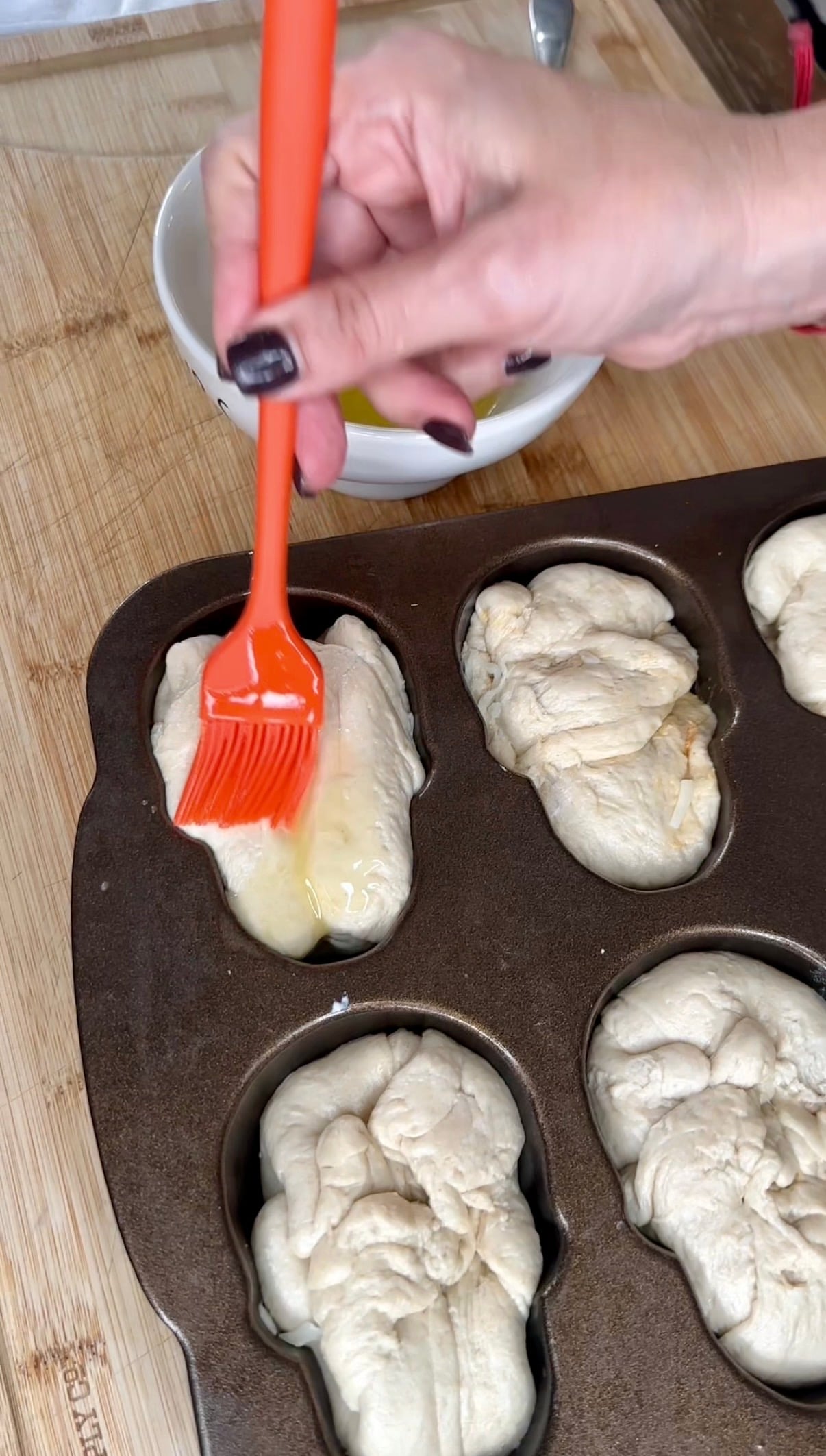 A hand uses an orange silicone brush to spread egg wash onto unbaked rolls in a muffin tin.