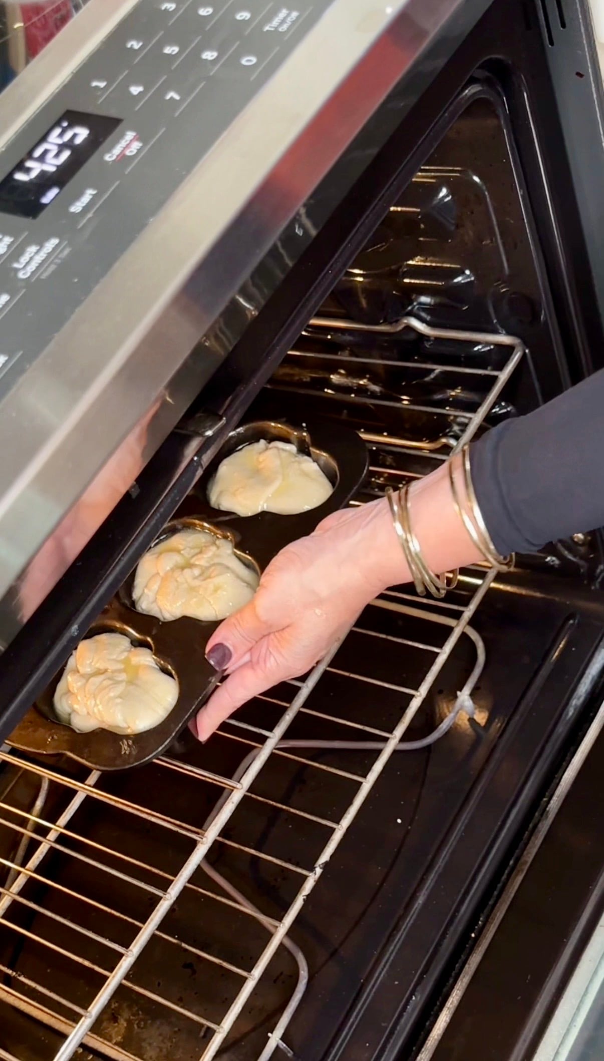 A hand places a muffin tin filled with three pieces of dough into an oven set to 425 degrees.
