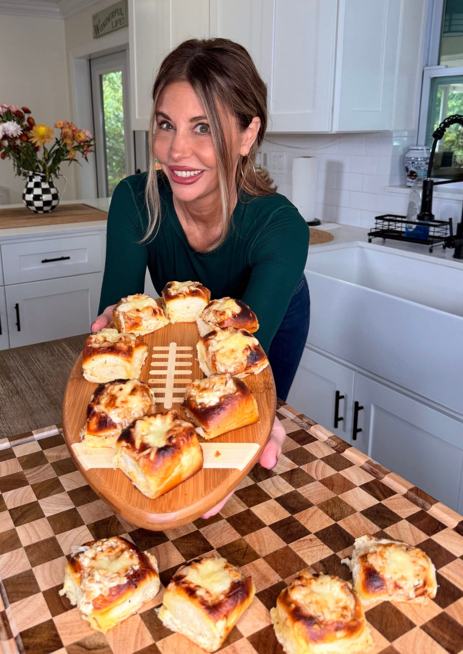 A woman in a green top smiles while holding a football-shaped tray of cheesy bread rolls in a kitchen. More bread rolls are on the wooden counter in front of her.