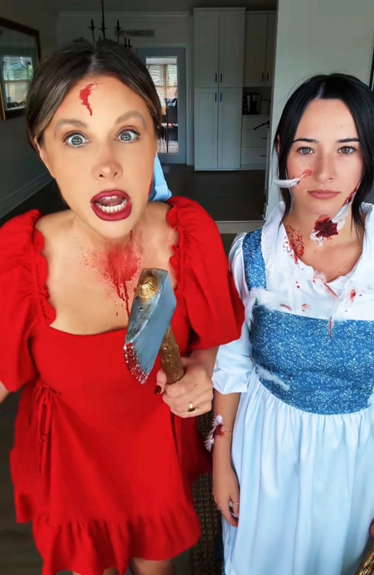 Two women in costume with fake blood and injuries: one dressed in a red dress holding a bloody axe, and the other dressed as Belle from Beauty and the Beast with feathers and blood on her clothing.