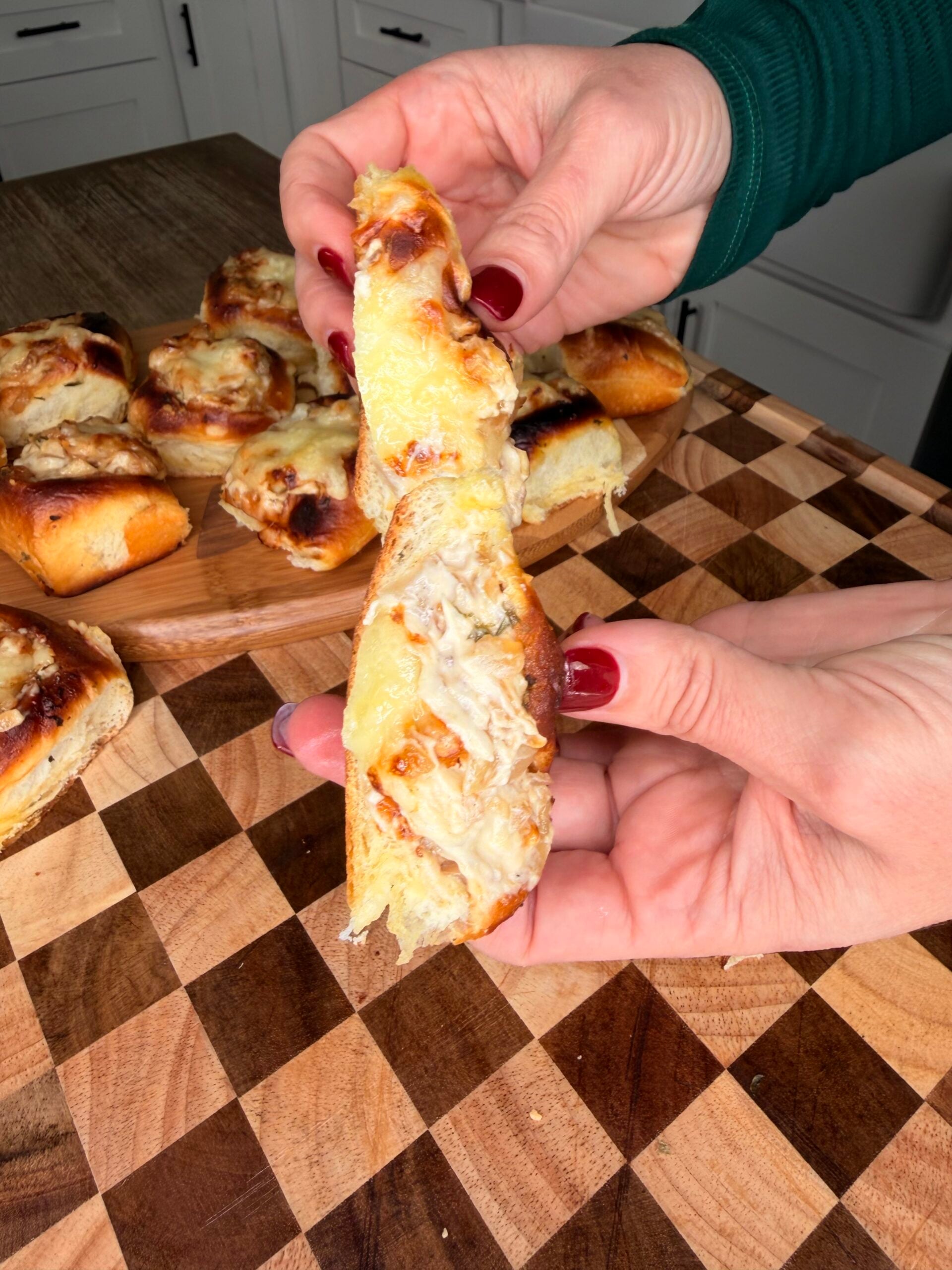 A person with red nail polish pulls apart a cheesy, baked bread roll, revealing a gooey cheese stretch, with more rolls on a wooden tray in the background.