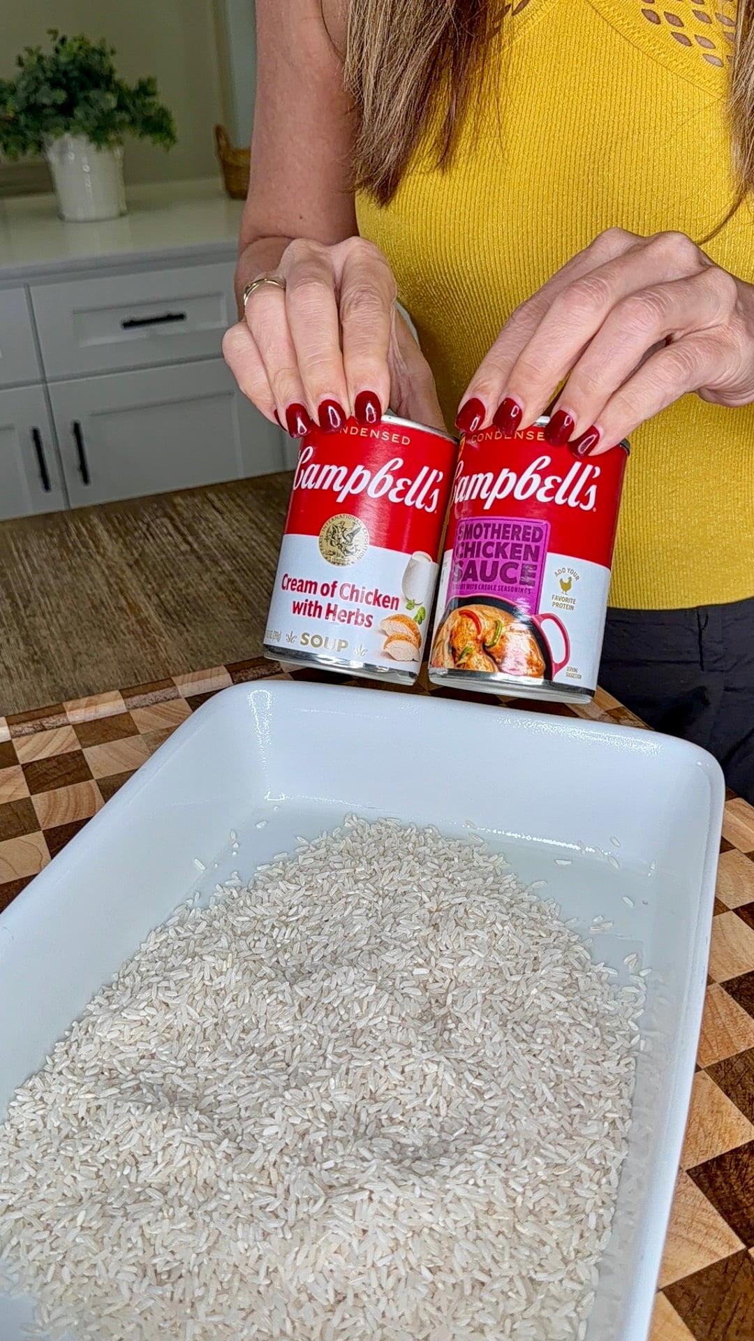 A person holds two cans of Campbells soupโCream of Chicken with Herbs and Smothered Chicken Sauceโabove a white baking dish filled with dry white rice.