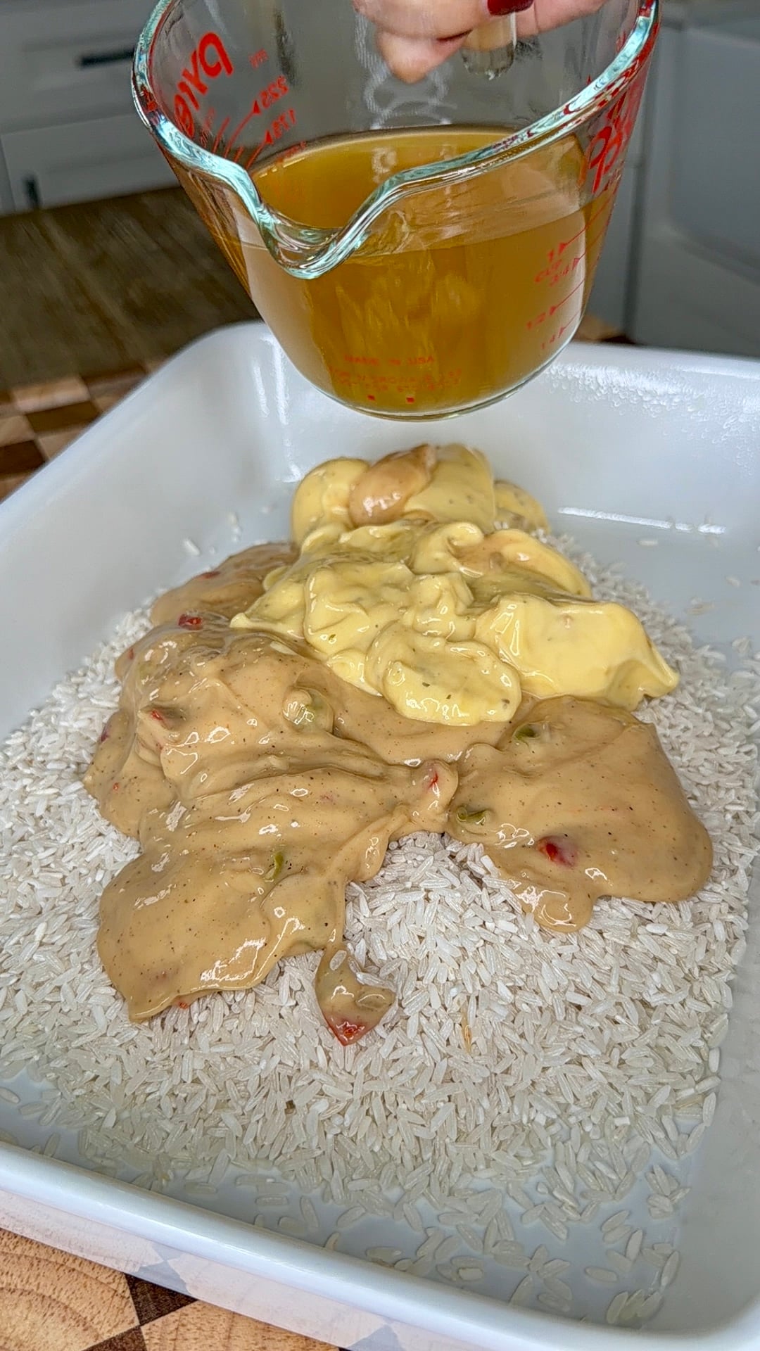 A measuring cup pours broth onto piles of raw white rice, cream of mushroom soup, and creamy cheese sauce in a baking dish.
