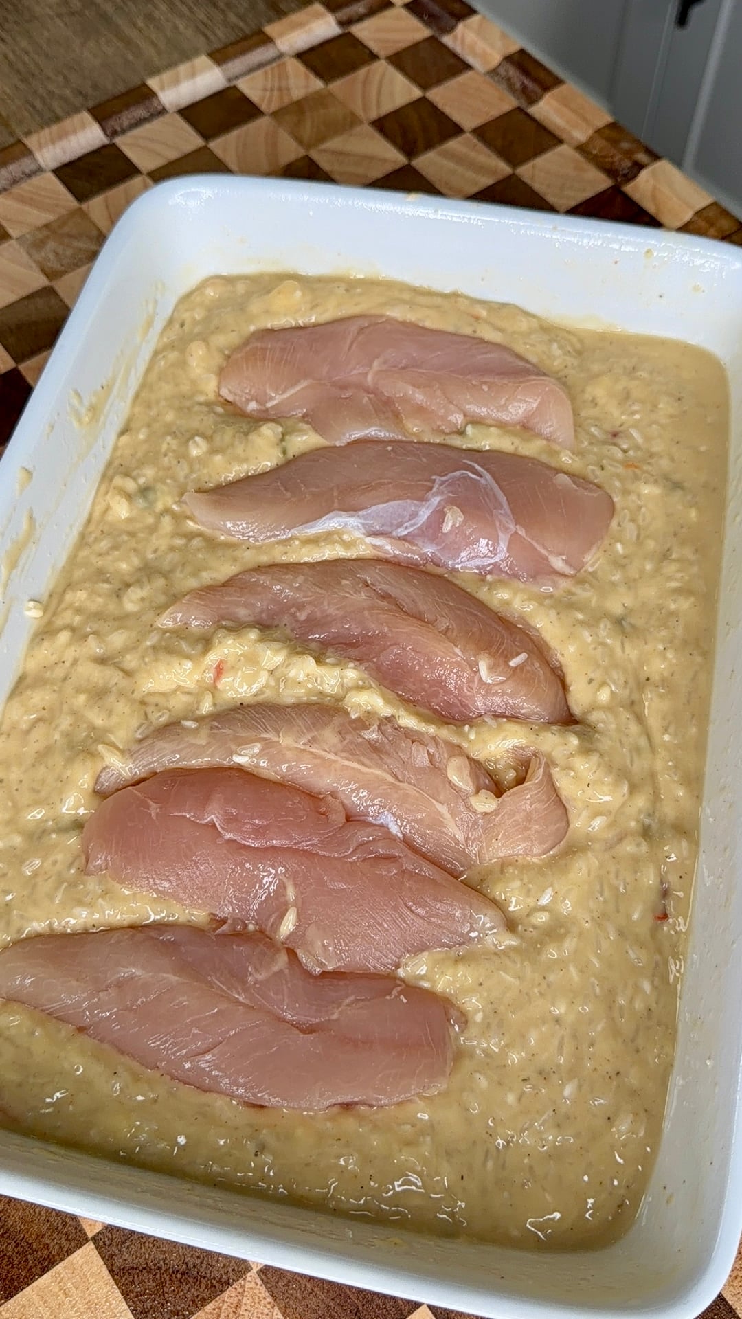 Five raw chicken breasts arranged in a row atop a creamy rice or casserole mixture in a white baking dish.