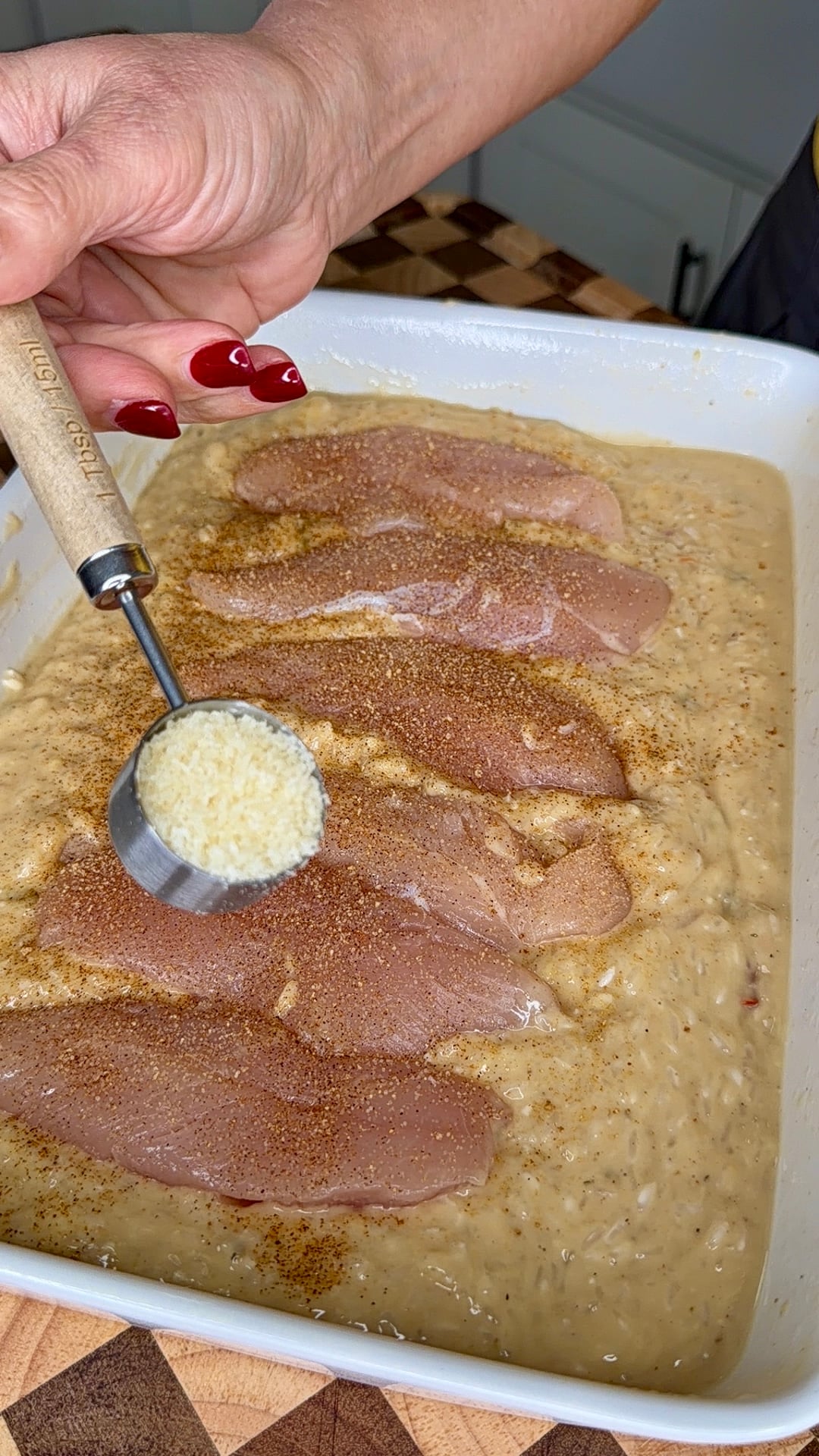 A hand uses a tablespoon measuring scoop to sprinkle grated cheese over raw chicken breasts resting in a baking dish of creamy sauce.