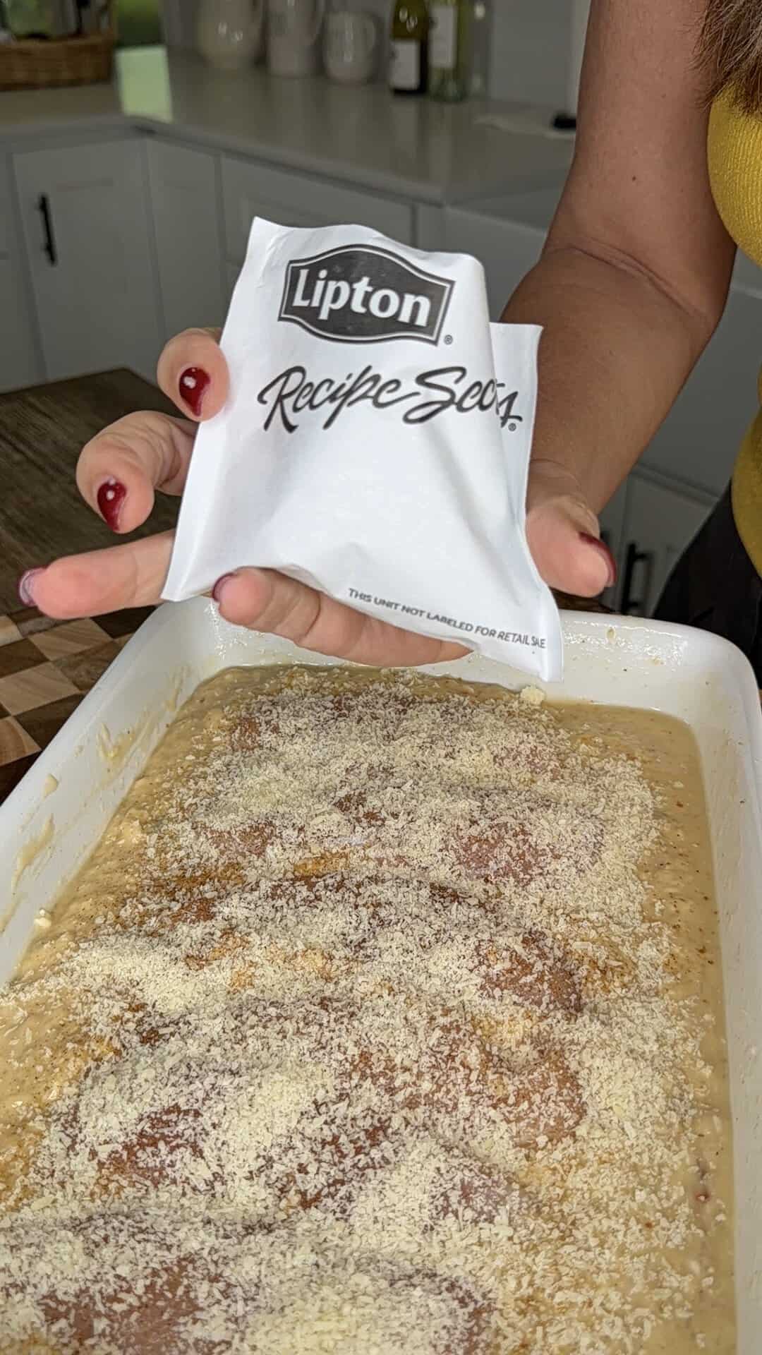 A person sprinkles a packet of Lipton Recipe Secrets mix over raw chicken breasts in a baking dish.