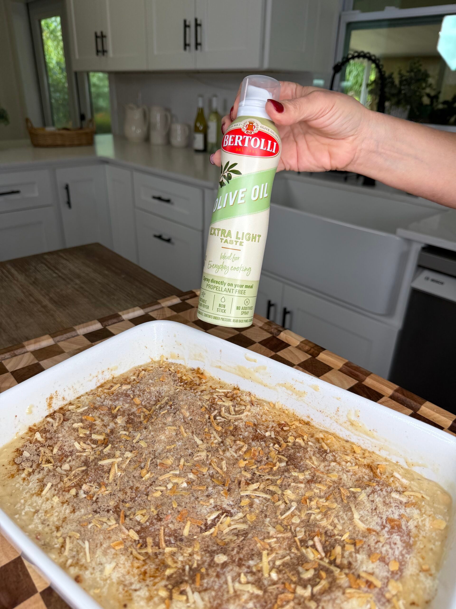 A hand holds a can of Bertolli Extra Light olive oil spray over a casserole dish filled with an unbaked, breadcrumb-topped meal.