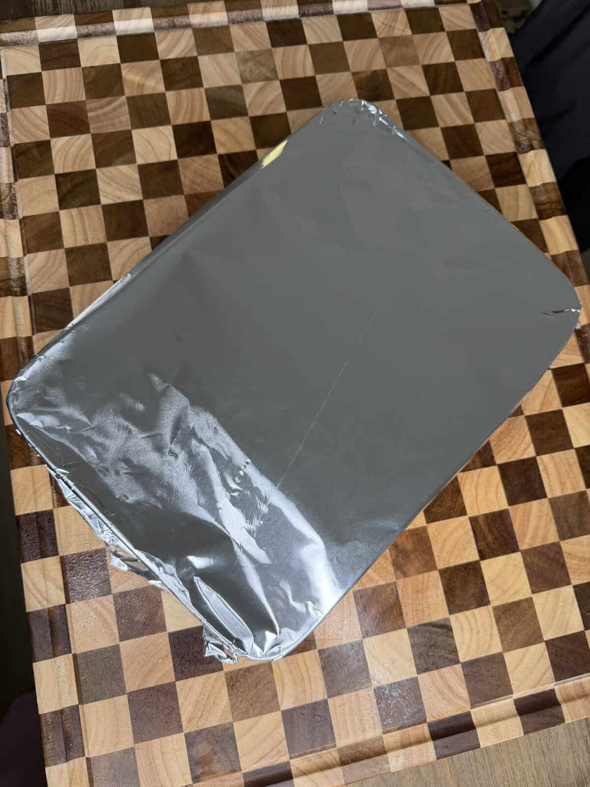A rectangular container covered in silver foil, sitting on a wooden checkerboard cutting board.