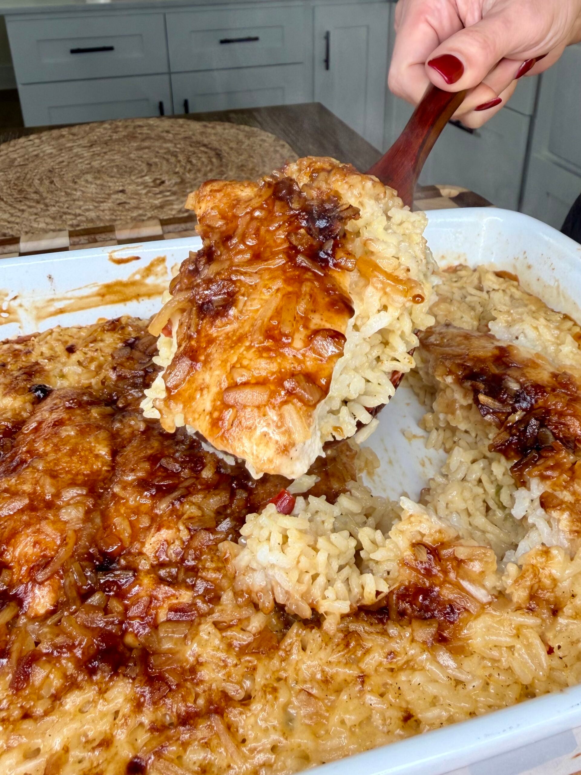A hand holding a wooden spoon scoops a serving of baked chicken and rice with a browned, caramelized onion topping from a white casserole dish.