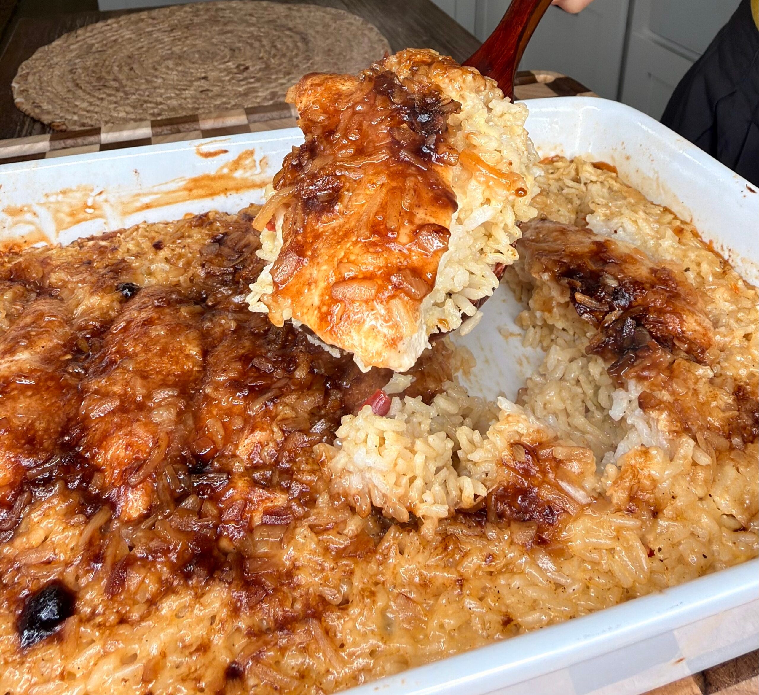 A hand holds a wooden spoon lifting a portion of baked rice with a caramelized top from a white casserole dish.