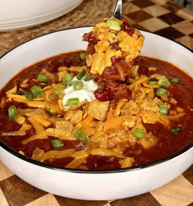 A spoonful of chili topped with shredded cheddar, tortilla chips, sour cream, and sliced green onions being lifted from a bowl.