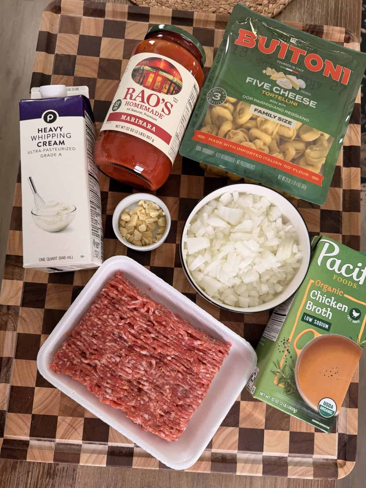 Ingredients for a meal including a carton of heavy cream, a jar of Raos marinara sauce, a bag of Buitoni five cheese tortellini, a bowl of chopped onions, a small bowl of minced garlic, raw ground meat, and a carton of organic chicken broth.