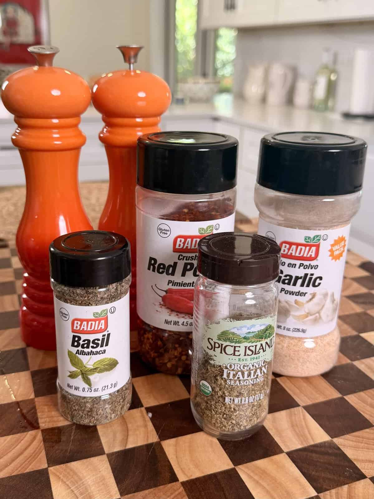 Four spice jars labeled Badia Basil, Badia Crushed Red Pepper, Spice Island Organic Italian Seasoning, and Badia Garlic Powder, arranged in front of two orange pepper mills on a checkered wooden board.