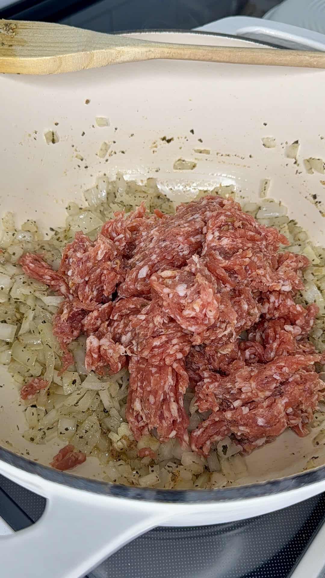 Ground meat piled on top of sautรฉed chopped onions in a white enameled pot with a wooden spoon resting across the top.