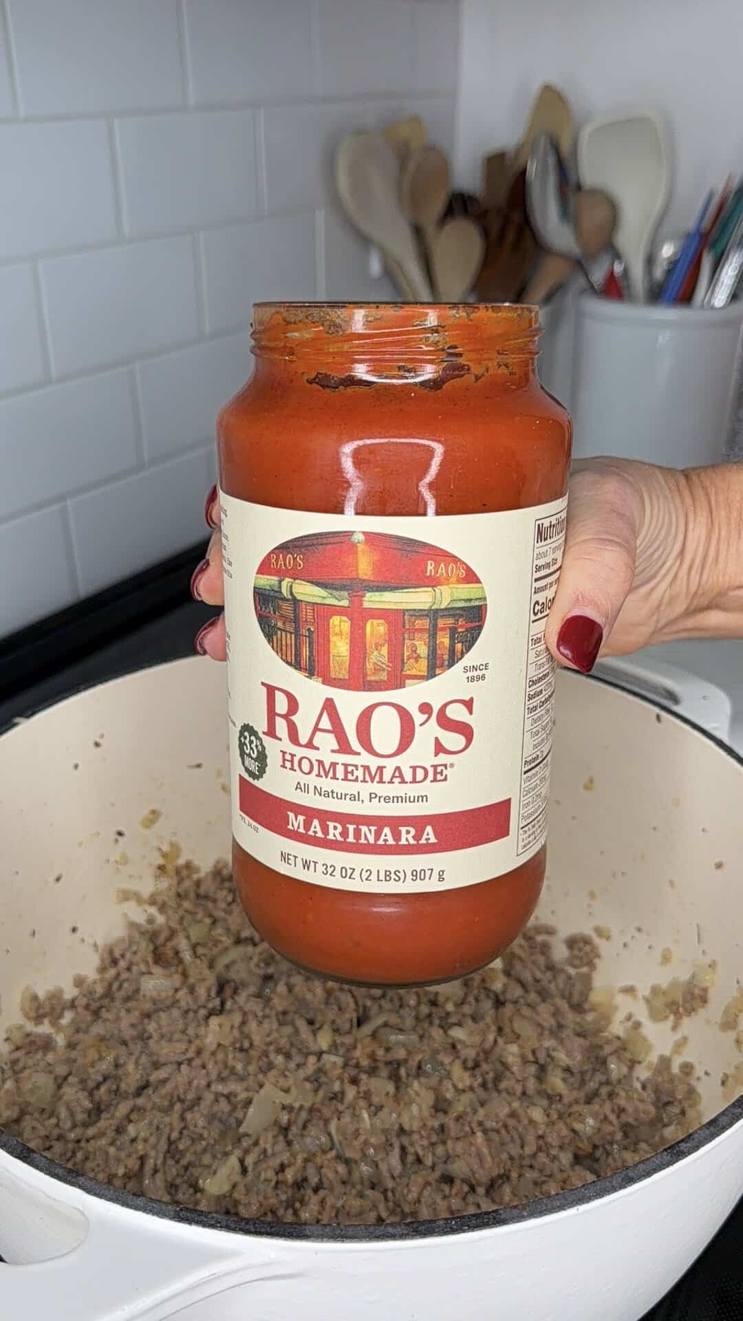A hand holds a jar of Raos Homemade Marinara sauce over a pot of cooked ground meat.