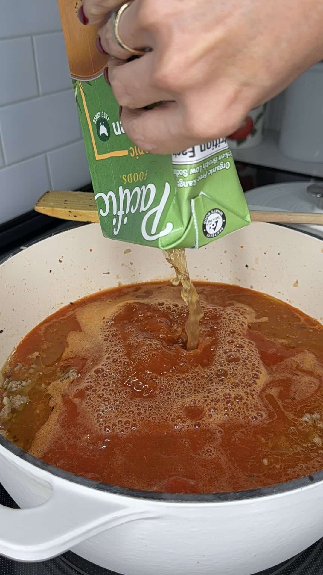 A hand pours Pacific Foods organic chicken broth from a carton into a pot of red liquid.