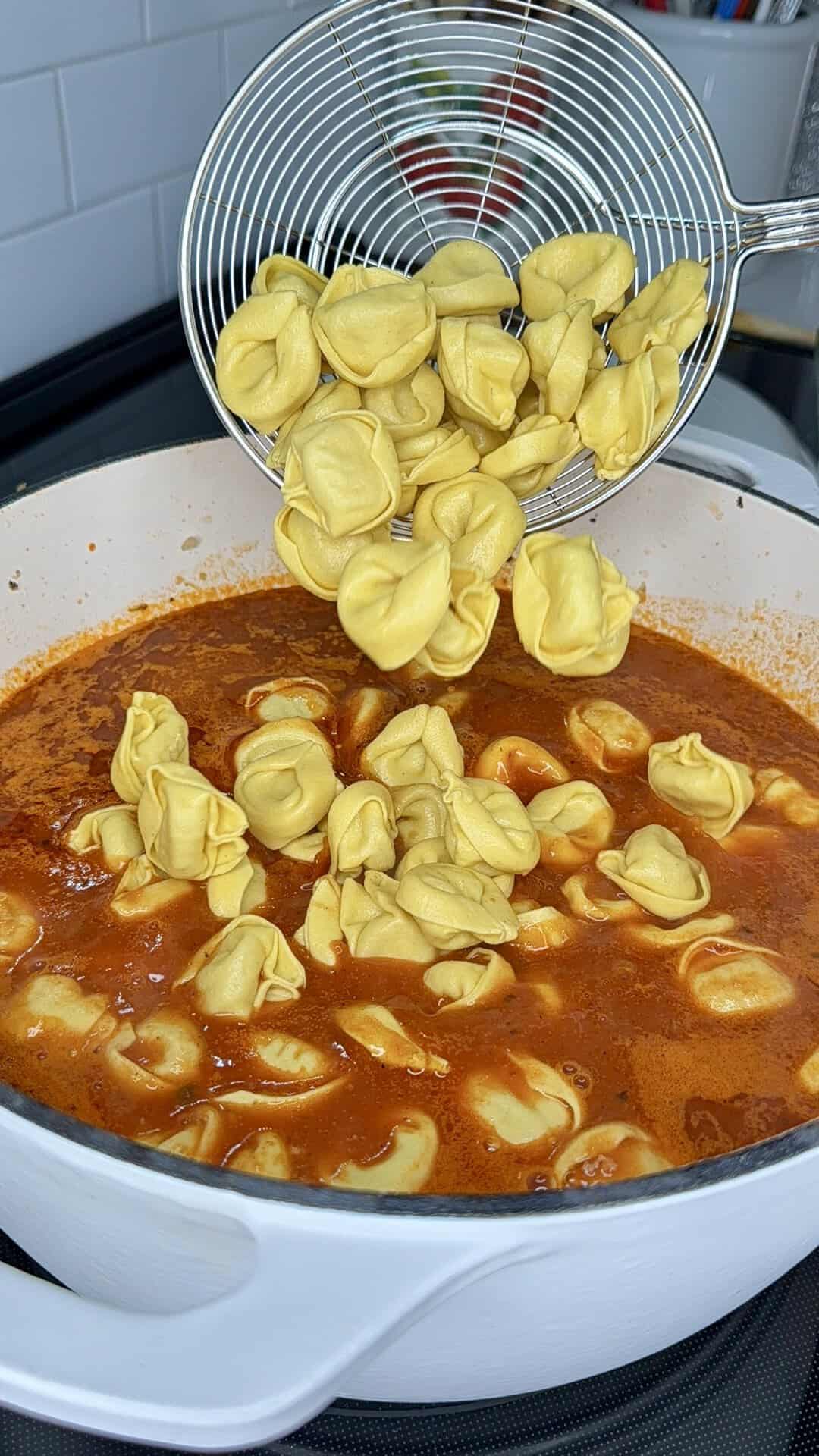 A metal strainer drops cooked tortellini into a large pot of red tomato sauce.
