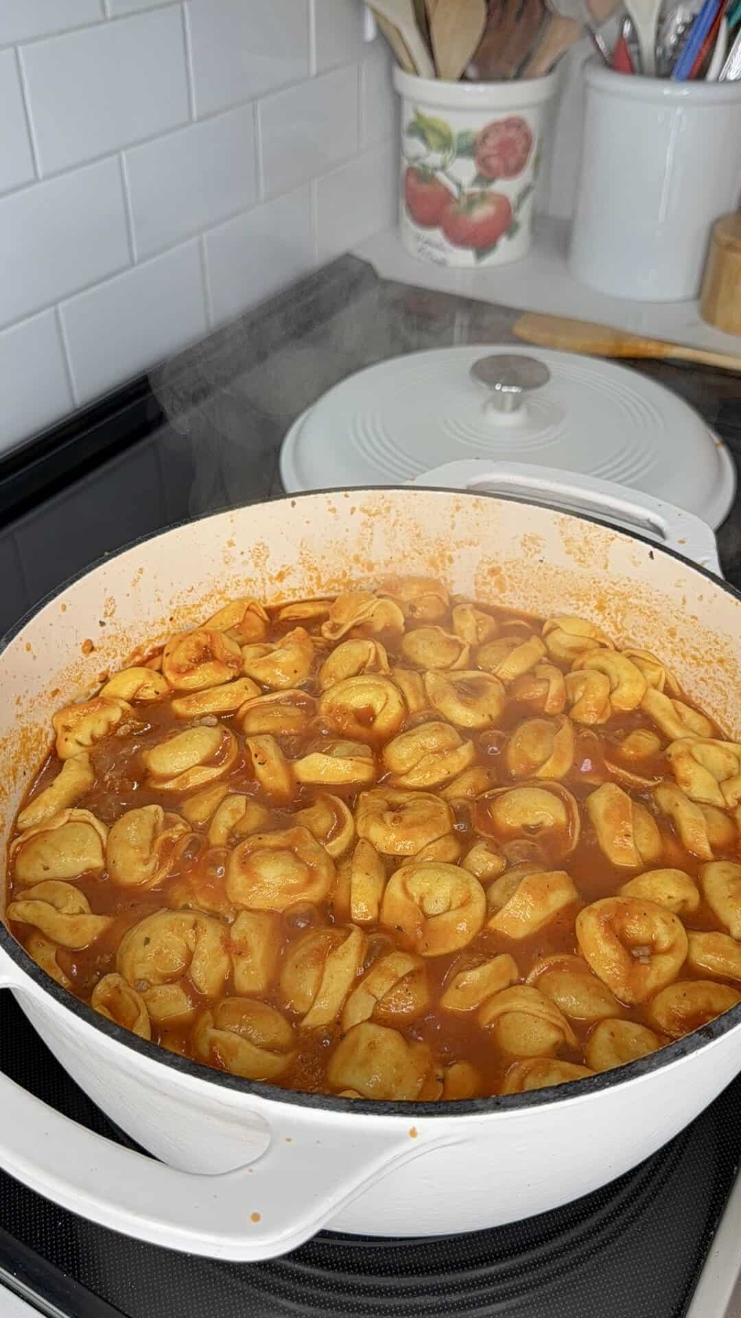Steaming tortellini simmering in a red tomato sauce in a large white pot on a stovetop.