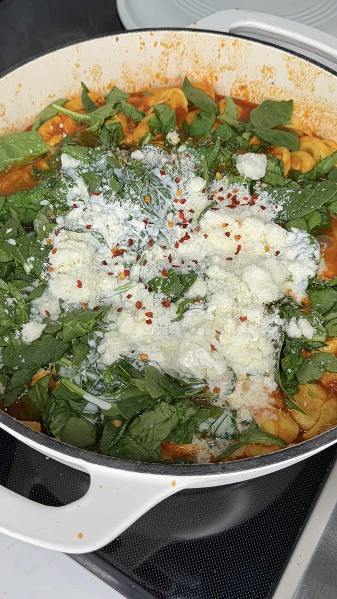 Tortellini in tomato sauce topped with fresh spinach, cream, grated cheese, and red pepper flakes in a white pot.