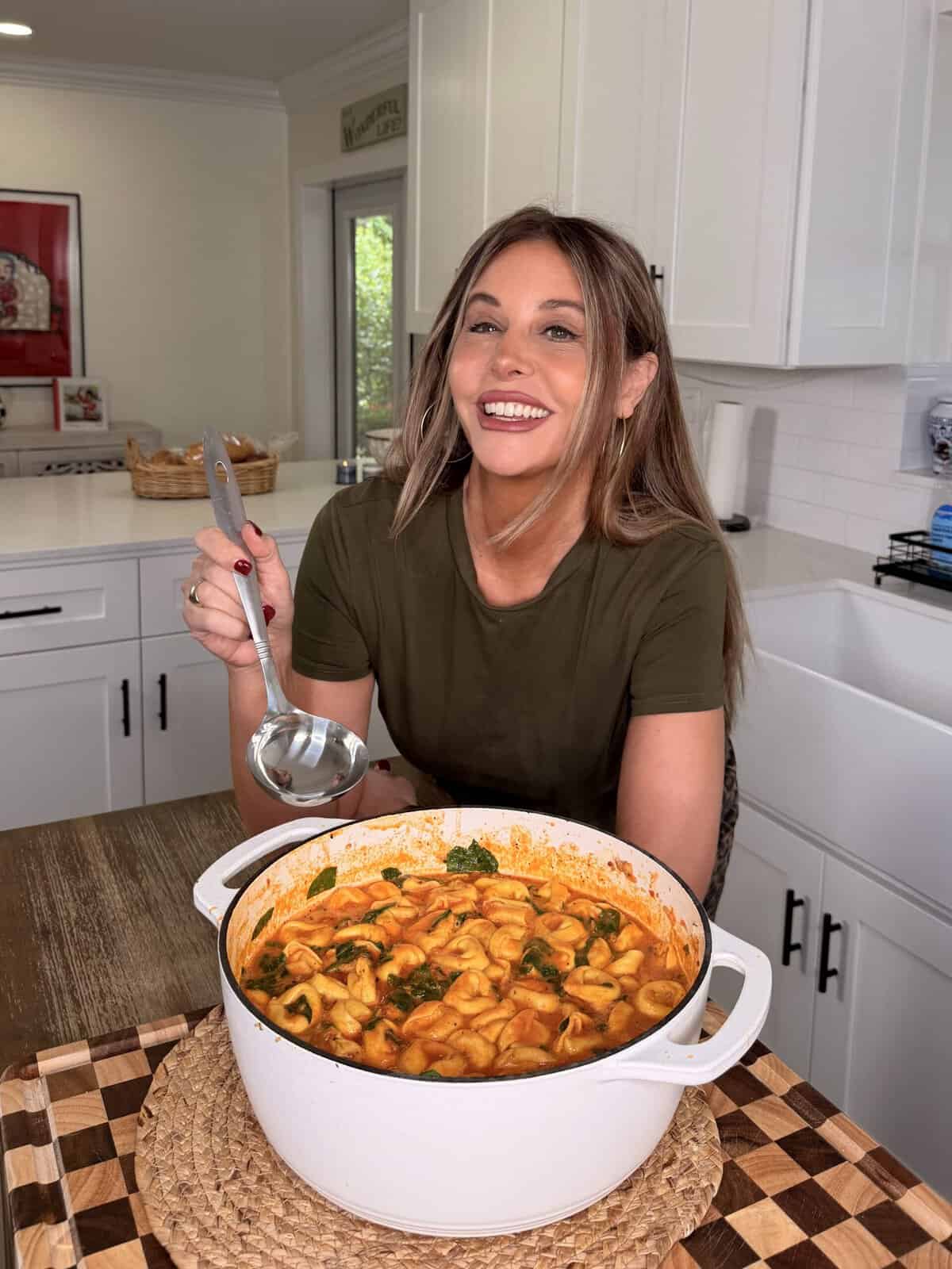 A woman in a kitchen holds a ladle over a white pot of tortellini soup with spinach.