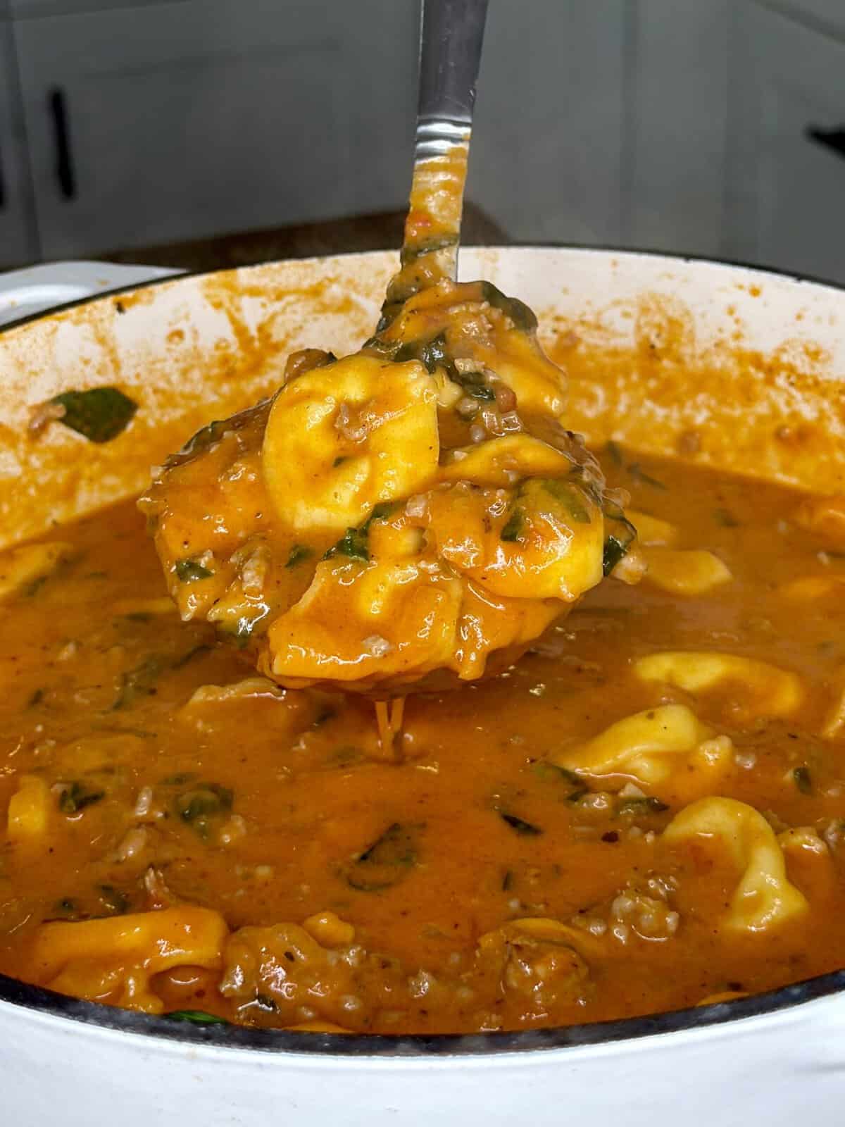 A ladle lifts creamy tomato soup with tortellini, ground meat, and spinach from a large pot.