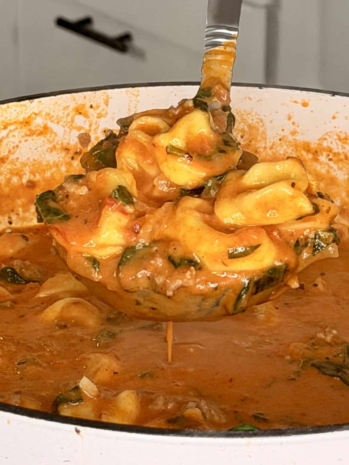 A ladle lifting creamy tomato tortellini soup with spinach from a pot.