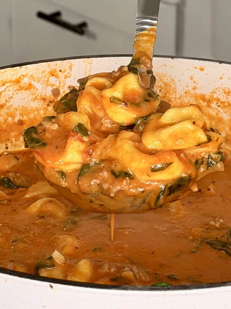 A ladle lifting tortellini and spinach in a creamy tomato sauce from a pot.