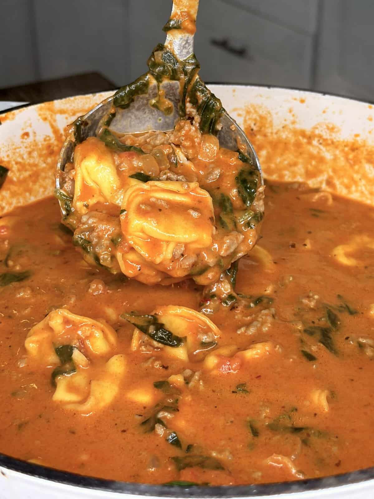 A ladle lifts creamy tomato soup with tortellini, ground meat, and spinach from a large pot.