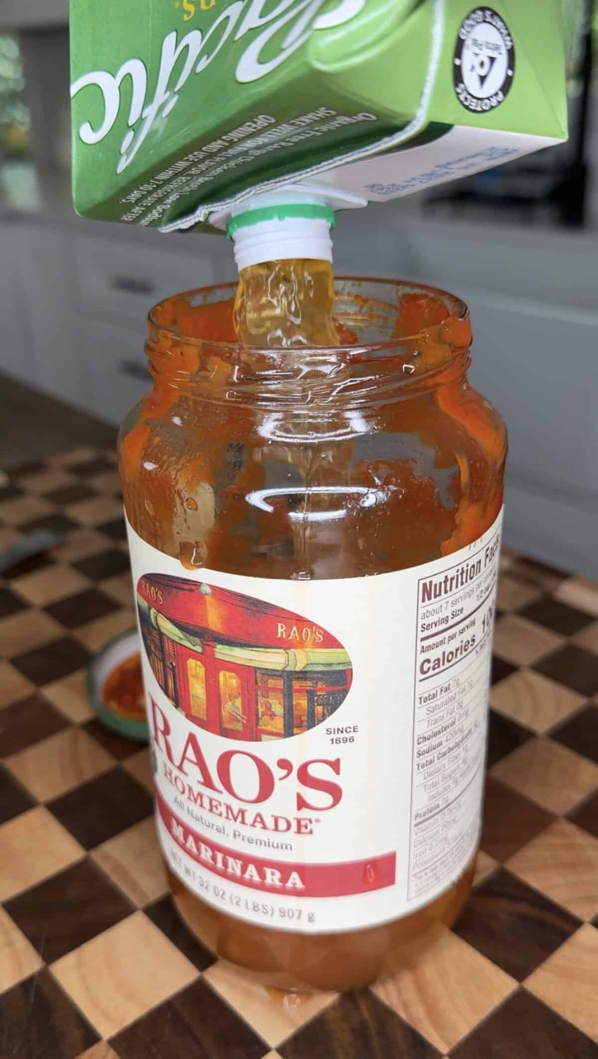 Broth from a green Pacific Foods carton being poured into a jar of Raos Homemade marinara sauce.
